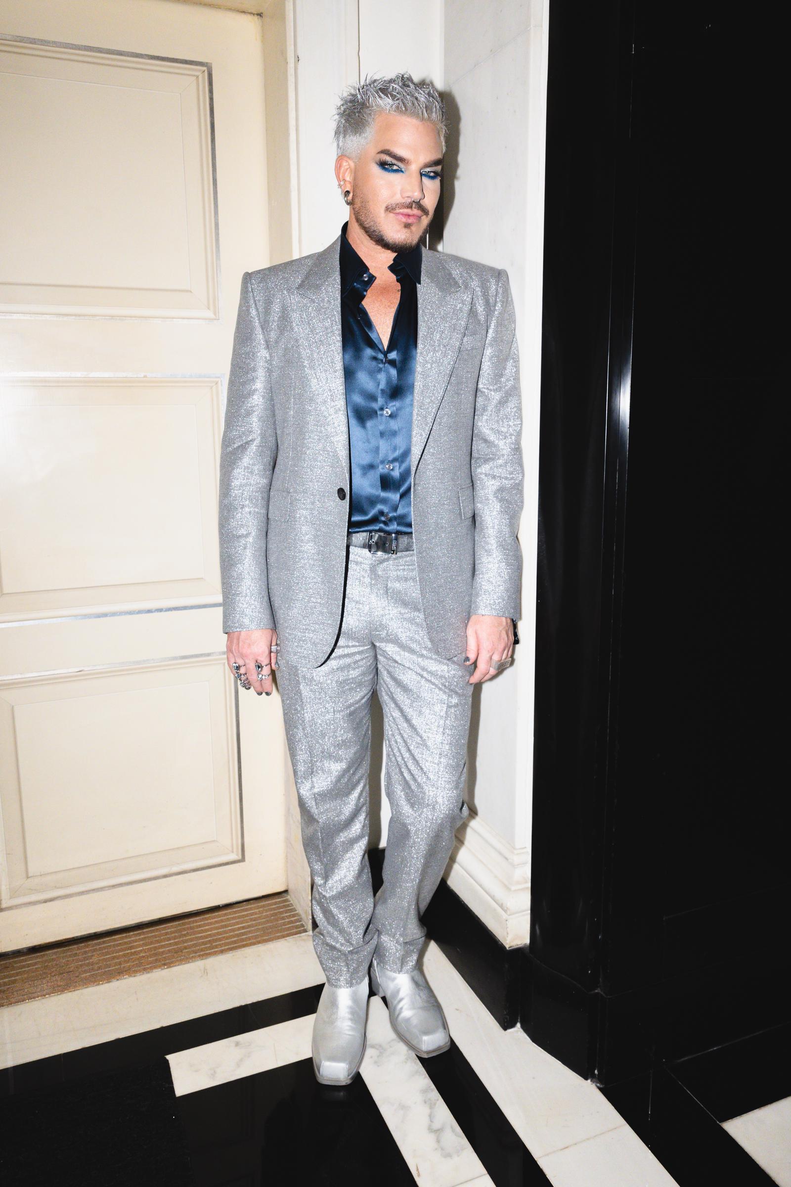 Adam Lambert poses against a cream wall in his silver suit and blue satin shirt, catching light with every detail. From the pointed shoes to the confident stance, the look feels bold yet polished, styled perfectly for a standout after-party moment.