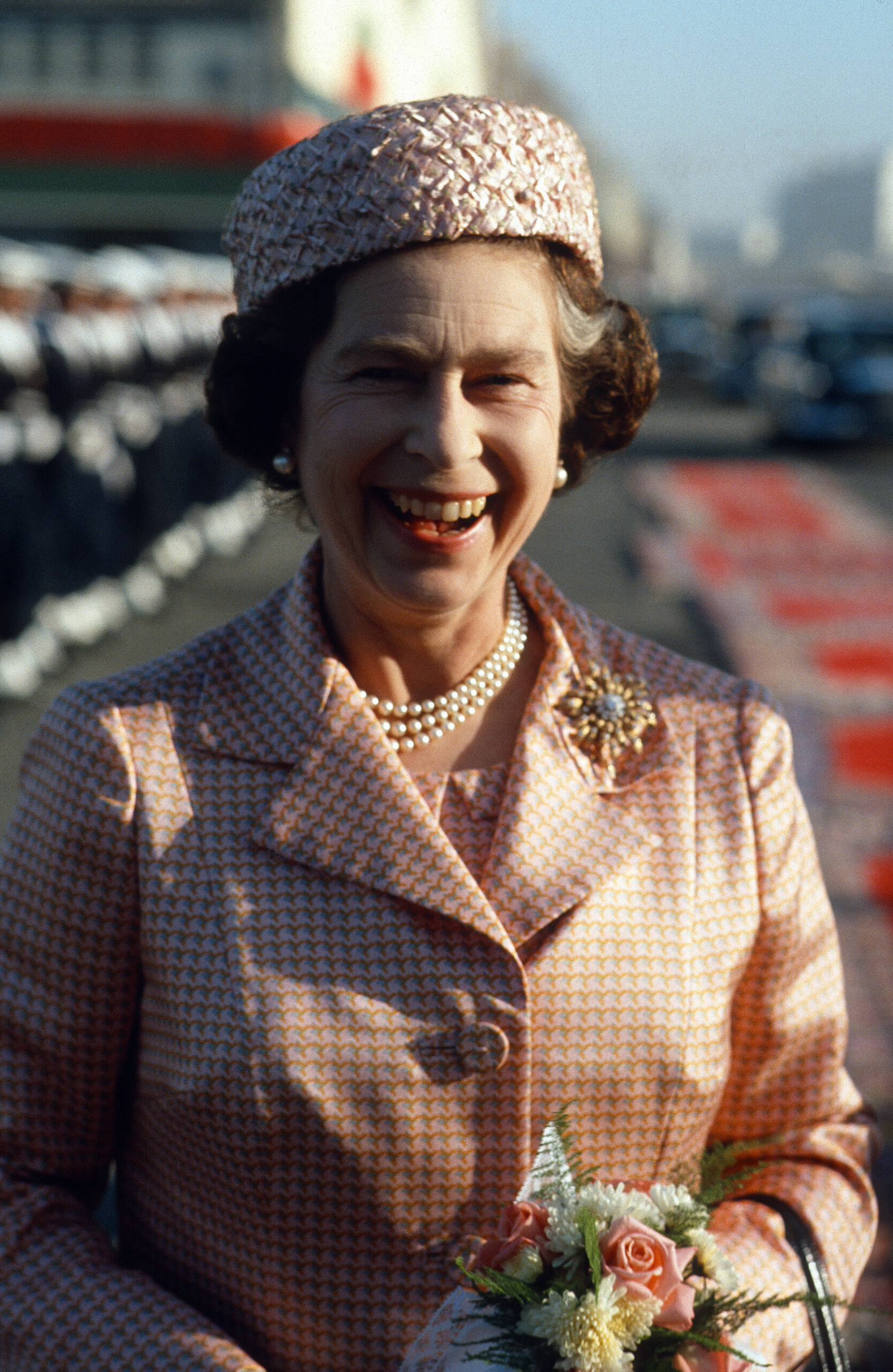Queen Elizabeth II shares a rare moment of unguarded laughter as she arrives in Casablanca on 27 October 1980. With a floral corsage pinned to her elegant patterned coat and a pillbox hat perched perfectly, the Queen radiates warmth on this unforgettable royal tour.