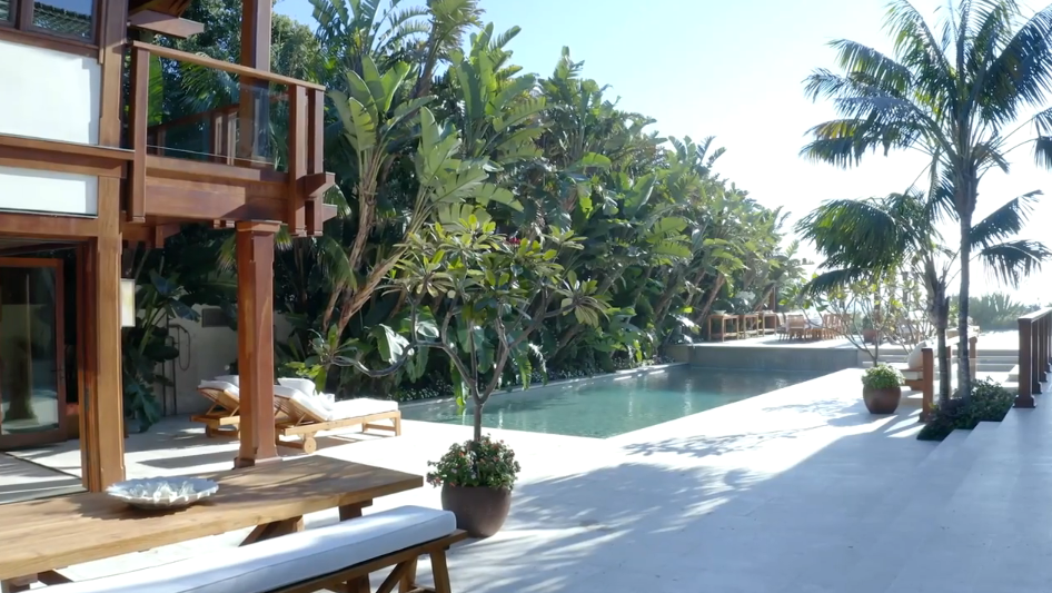 Surrounded by dense tropical greenery, a serene resort-style pool area features light stone decking, comfortable teak lounge chairs, and a shaded outdoor dining setup. Fragrant frangipani trees and soaring palm trees frame the poolside, creating a secluded oasis right outside the main dwelling. | Source: YouTube/Architectural Digest