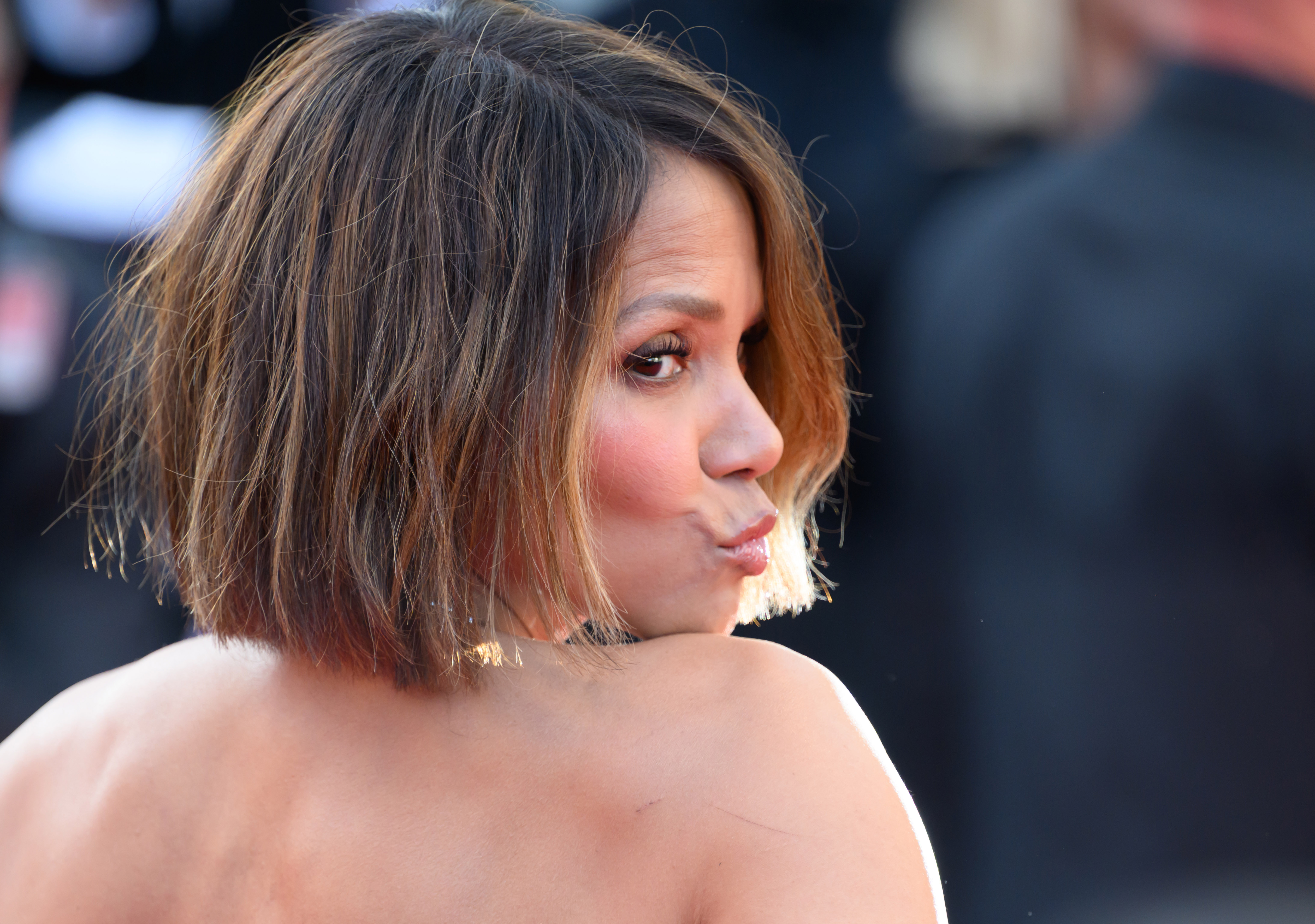 Halle Berry puckers her lips in a playful pose, glancing back over her shoulder with a cheeky expression as she engages with the cameras on the red carpet.