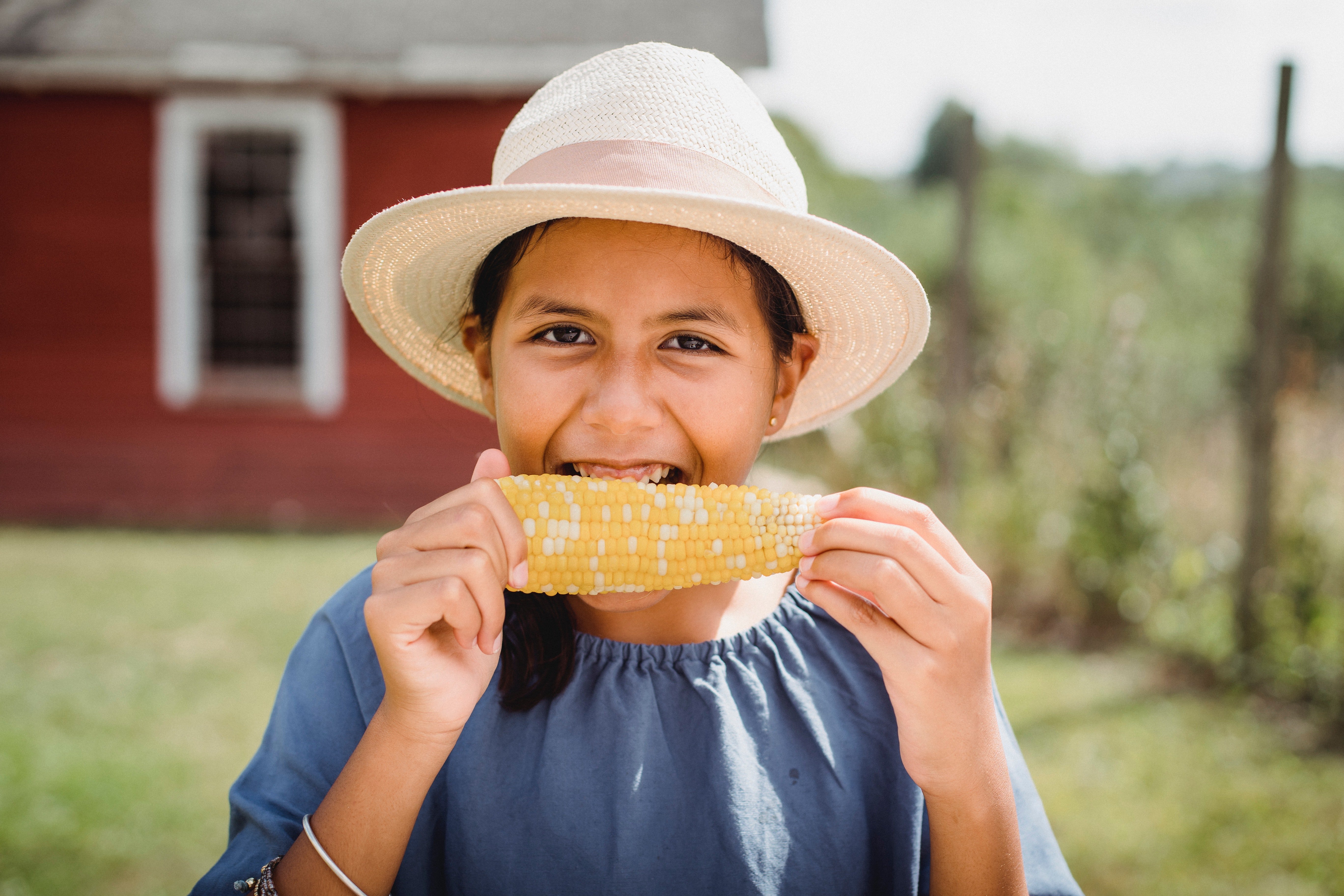 Pictured - A smiling young girl eating corn in the countryside wearing a blue top and a pink hat | Source: Pexels 
