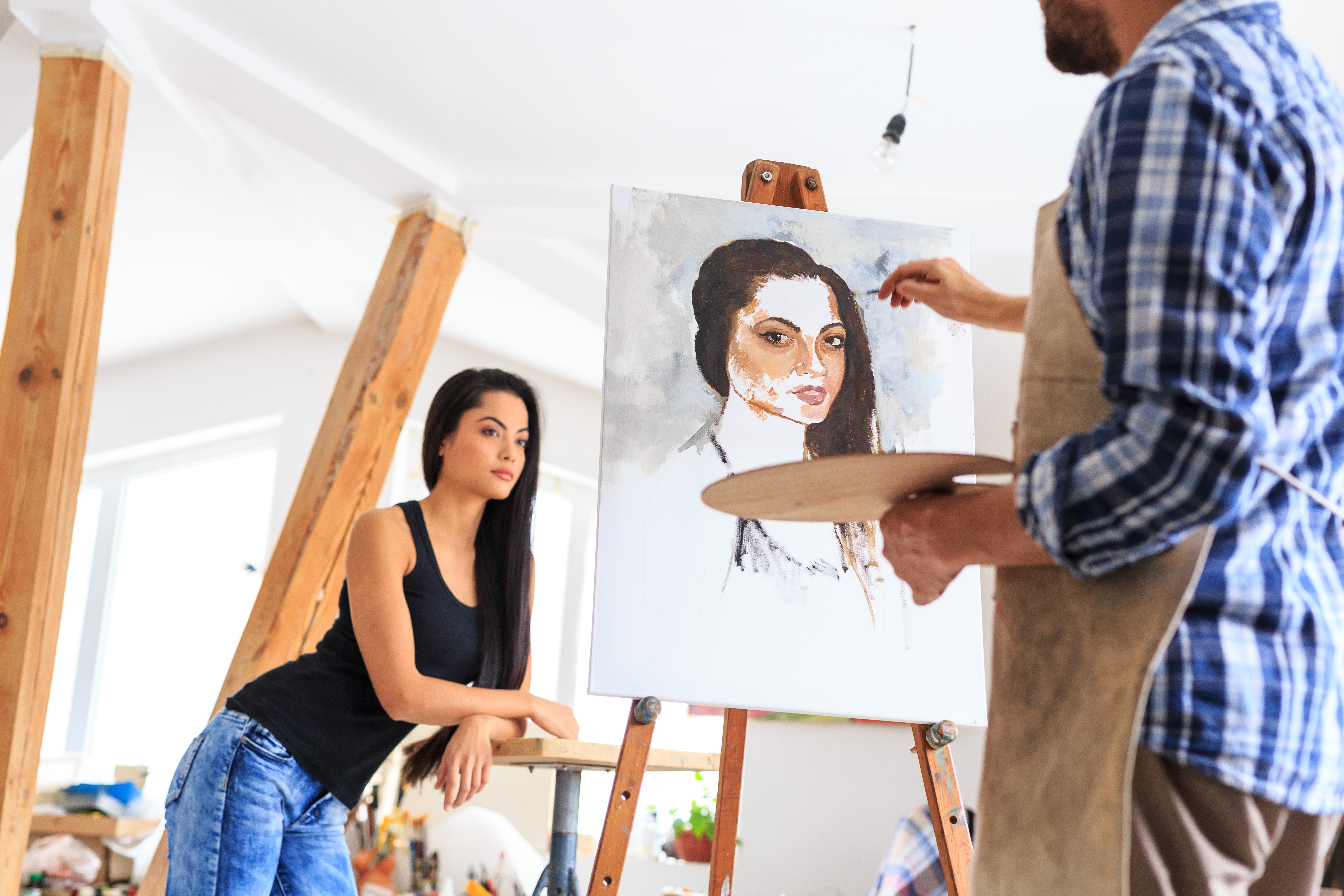 Rear view of young painter holding a color palette and making portrait of young woman in his art studio | Photo: Getty Images