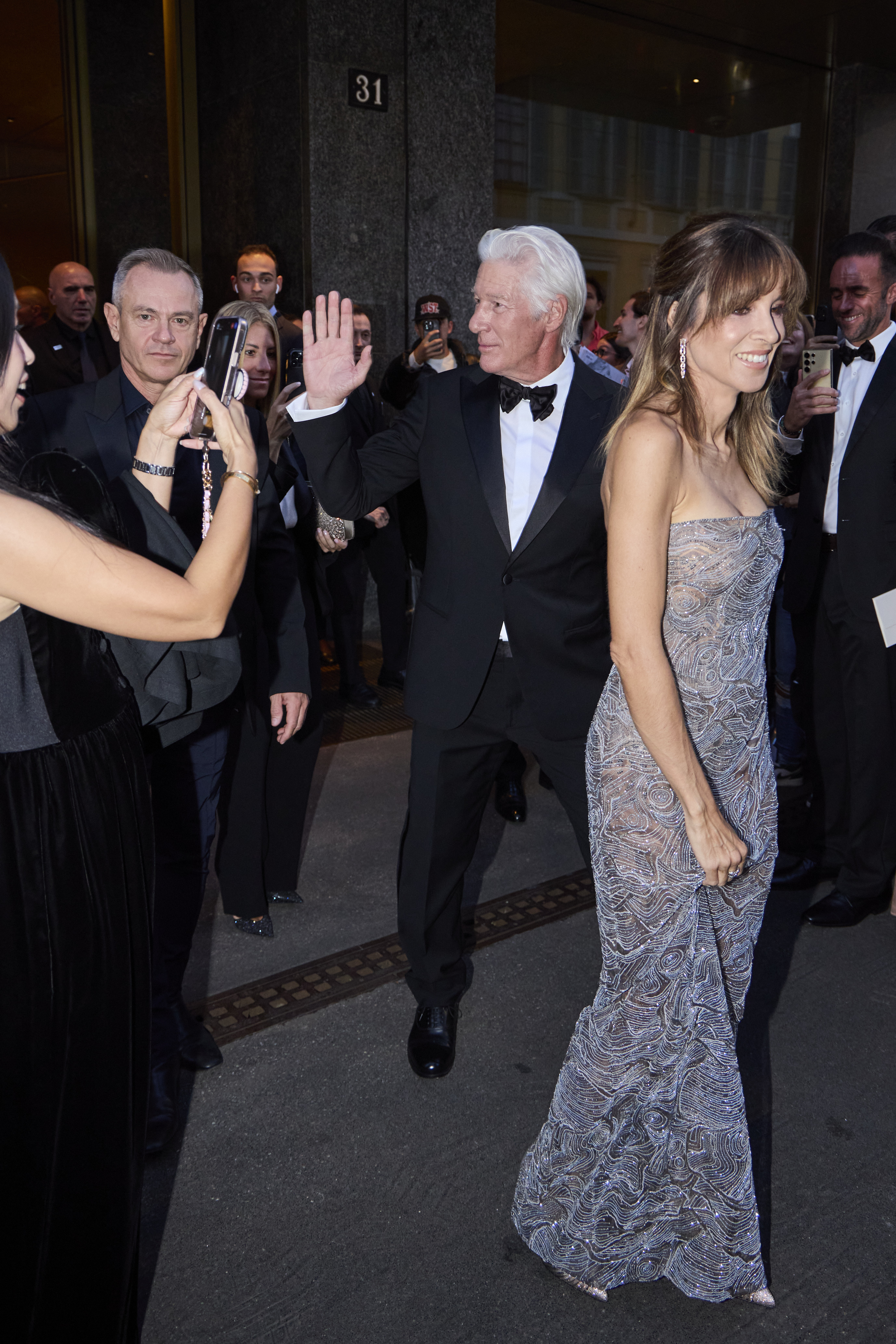Surrounded by flashing cameras, Richard Gere and Alejandra Silva make a grand entrance outside the Armani Hotel, smiling as fans rush to capture the moment.