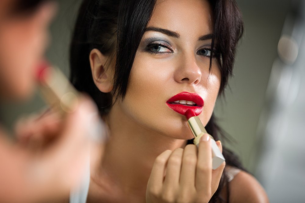 Mujer viéndose al espejo mientras se maquilla los labios de color rojo intenso. | Foto: Shutterstock