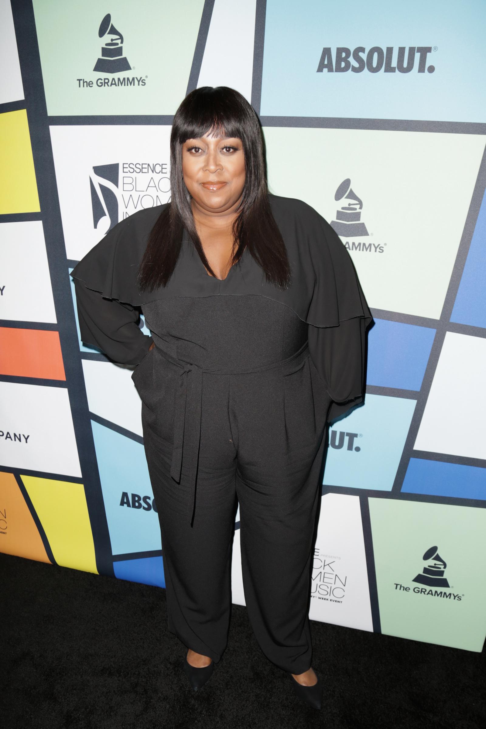 Loni Love poses at the Essence Black Women in Music event on February 9, wearing a flowing black ensemble with straight hair and a composed expression against the event's colorful backdrop.