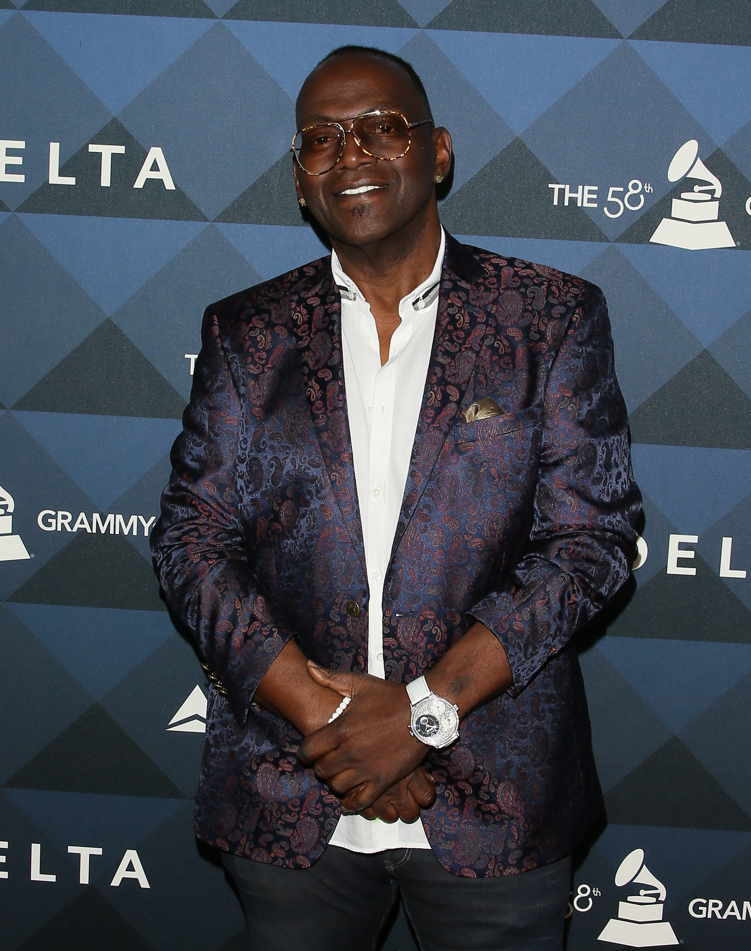 Wearing oversized tinted glasses and a patterned blazer, Randy Jackson poses with folded hands against a sleek GRAMMY Weekend backdrop.