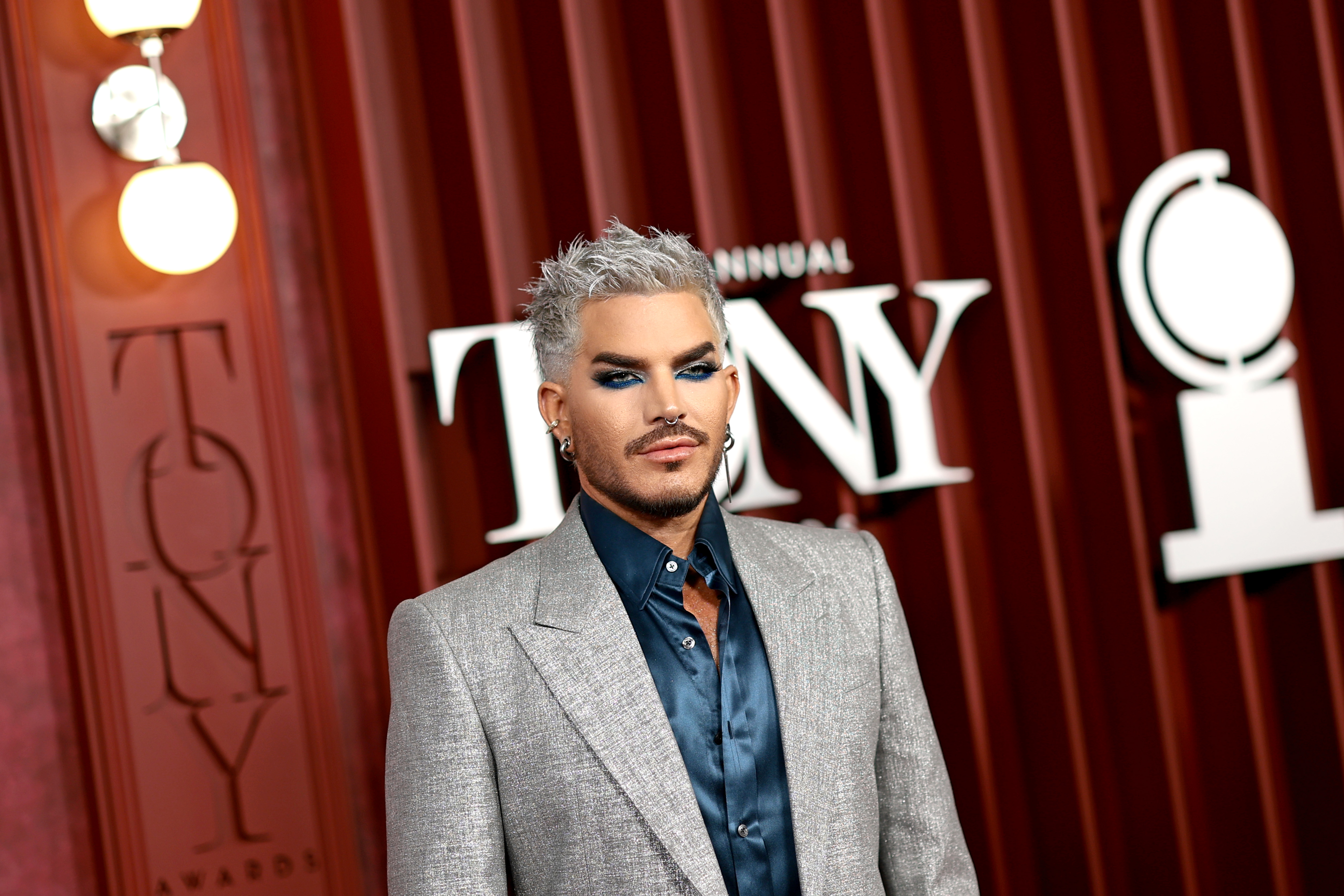 Striking a poised stance, Adam Lambert commands attention in a structured silver blazer and deep blue shirt. His frosty hair, metallic hues, and confident styling blend edge with refinement, elevating a moment of standout fashion expression.