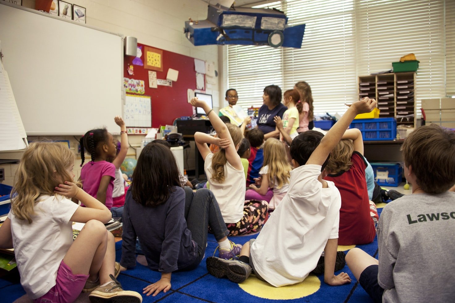 Niños sentados en el suelo de un aula de clases, levantan la mano. | Foto: Unsplash