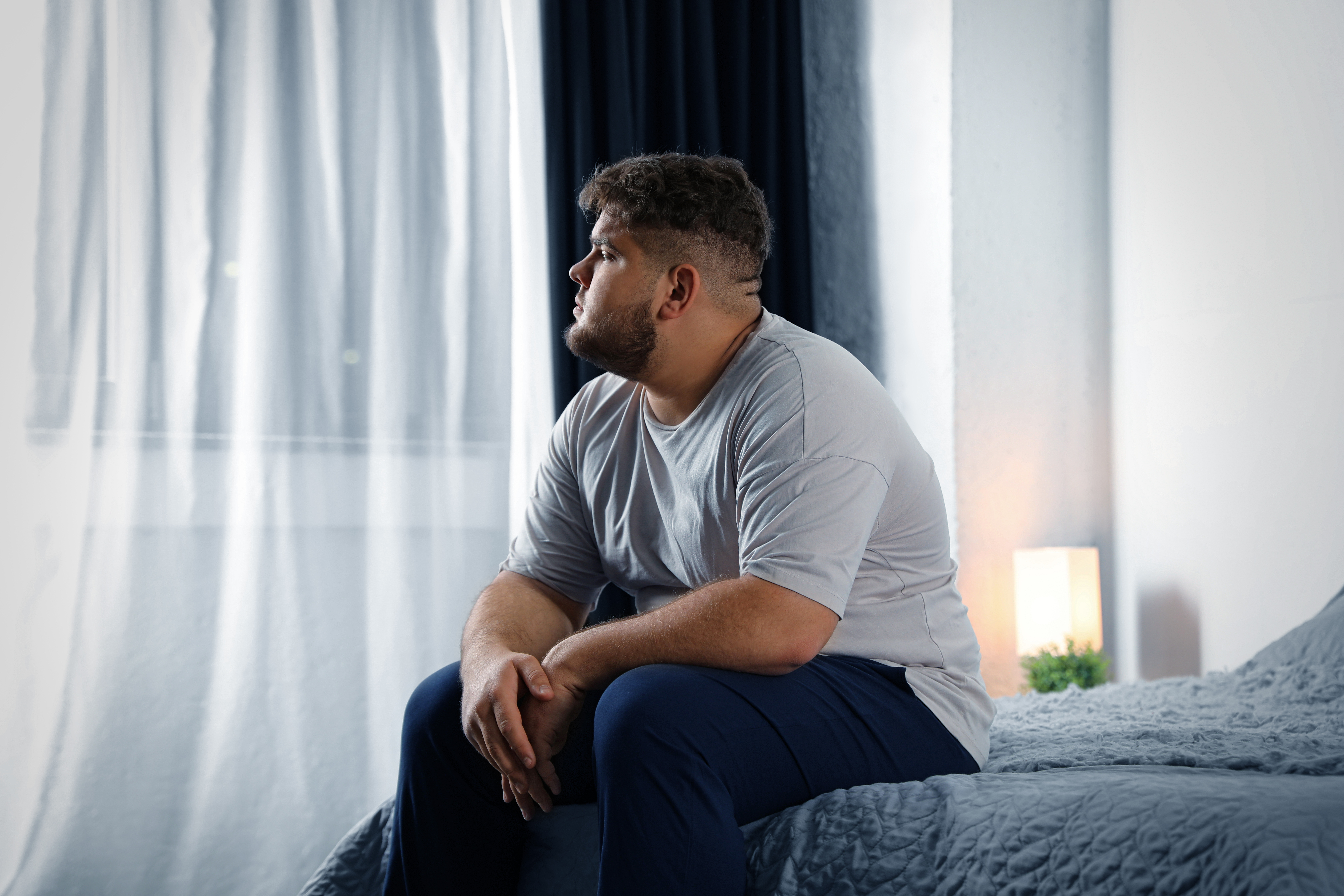 A depressed, overweight man is pictured sitting on his bed at home | Source: Shutterstock