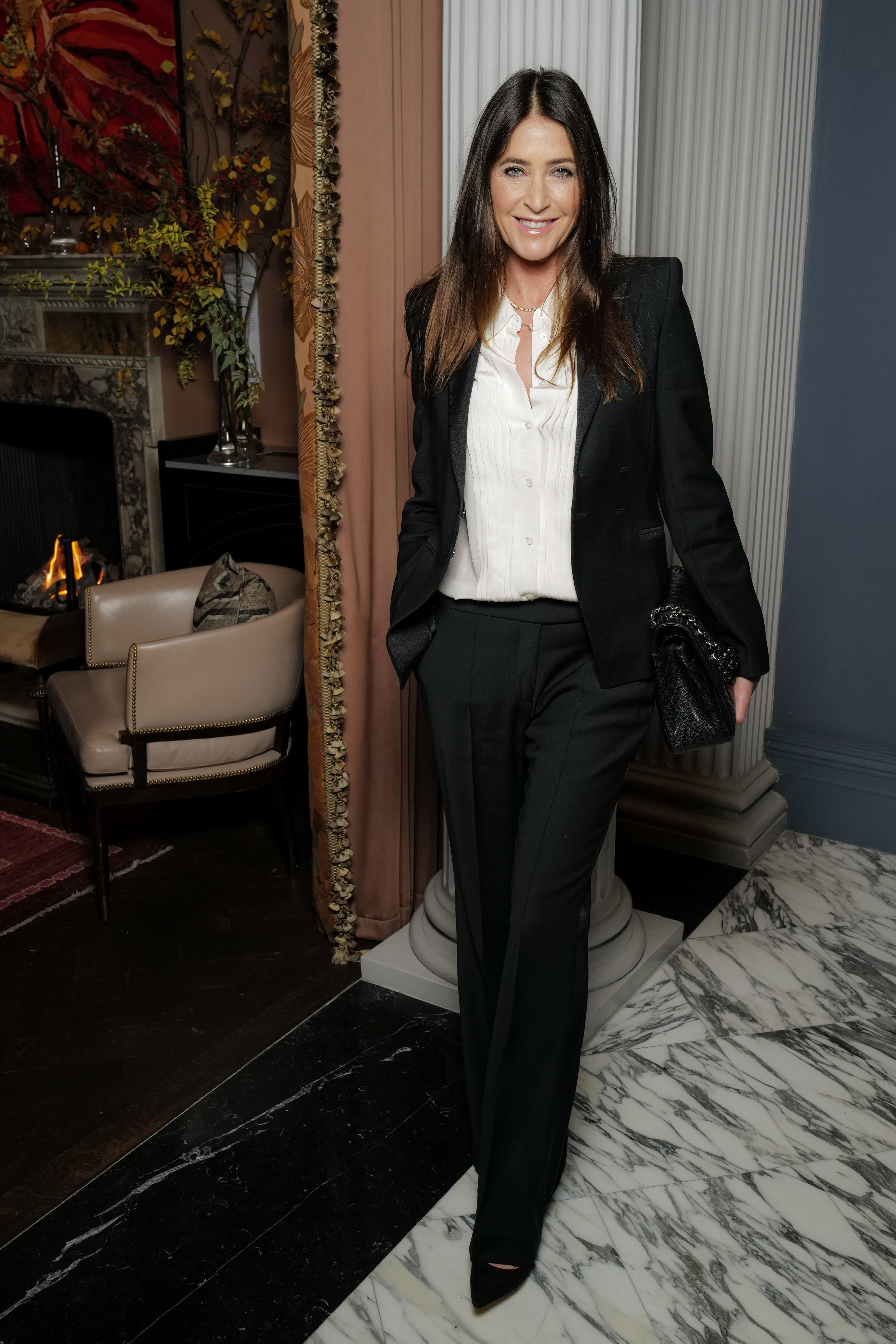 Estée Lauder and Harrods Dinner, 3 November 2025 — Inside an elegantly appointed room with a lit fireplace and marble floors, Snowdon wears a sharp black trouser suit over a white satin ruffle-front blouse, a quilted black leather bag tucked under her arm