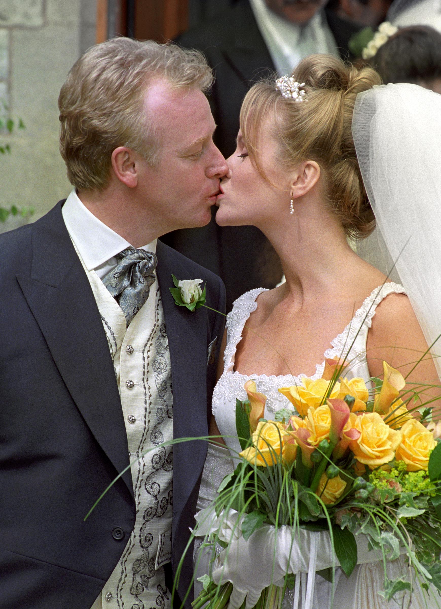 Les Dennis, dressed in a navy morning suit with an ornate cream waistcoat and white rose buttonhole, shares his first kiss with bride Amanda Holden outside the church in Bournemouth. Holden wore a white lace-trimmed gown with a pearl hair accessory and veil, clutching a bouquet of yellow roses and calla lilies.