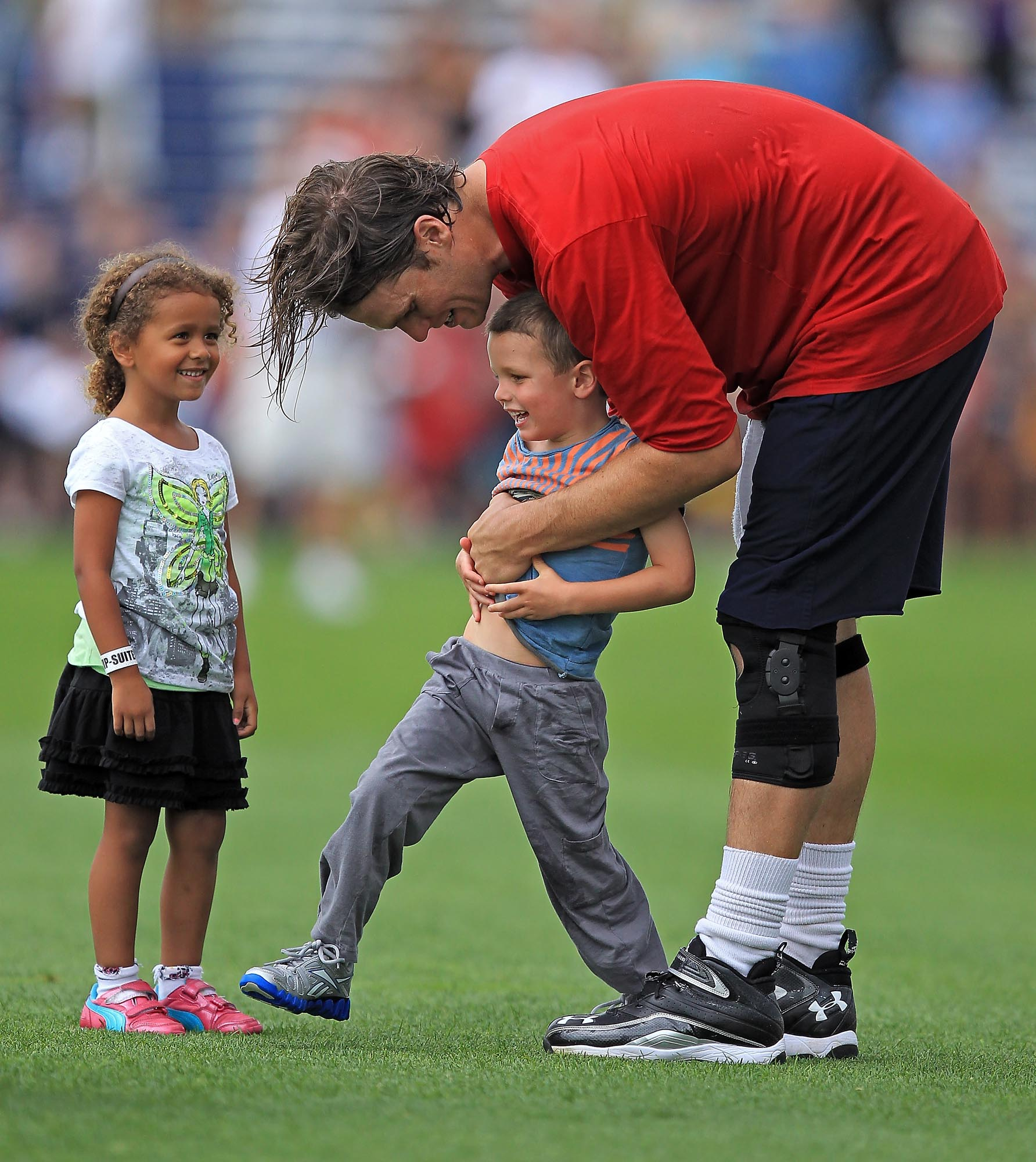 On the practice field, Tom Brady embraces Jack Moynahan with a laugh while his niece Jordan looks on, capturing a tender family moment after a Patriots practice session.
