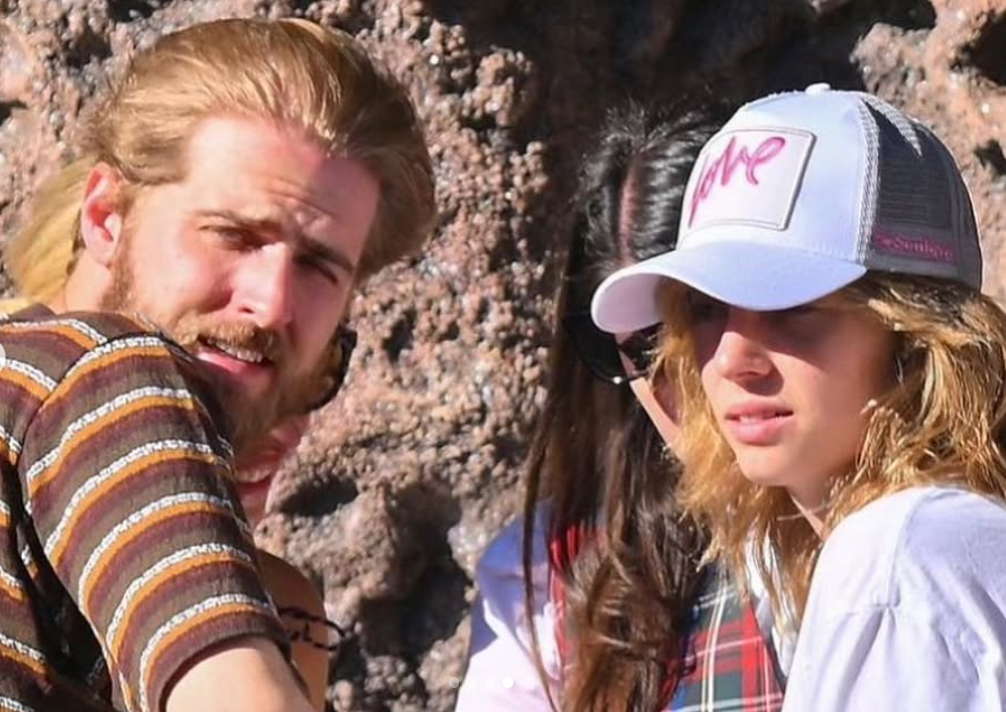 In bright sunlight against a rocky backdrop, Christian Hutson looks over his shoulder while Maya Hawke in a white trucker hat glances toward the camera. | Source: Instagram/maya_hawke