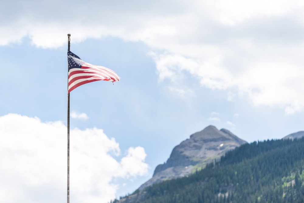 A flagpole with the American flag waving in the wind and mountain peak in summer. | Photo: Shutterstock
