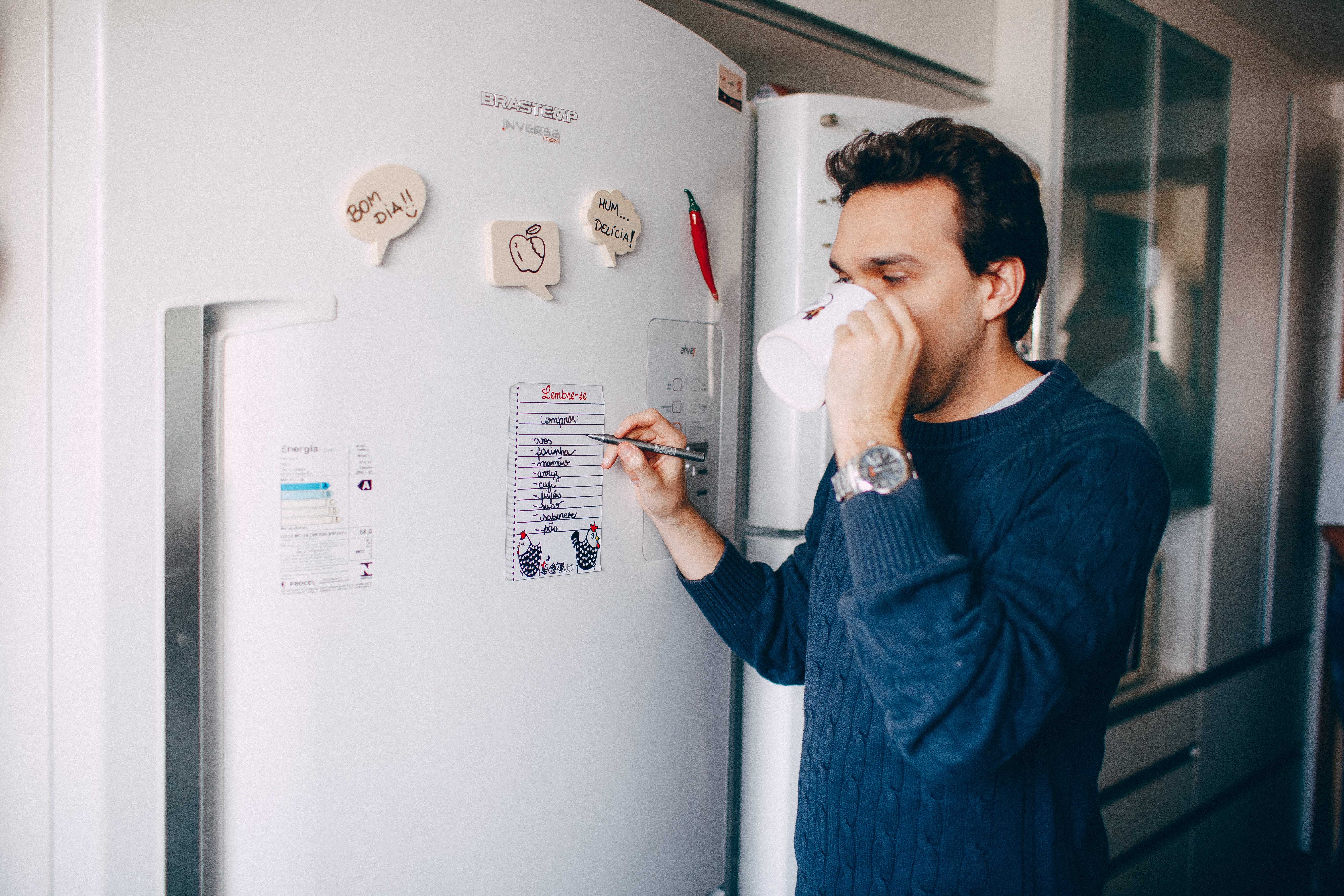 A man drinking from his mug while writing something on a paper stuck on a fridge | Source: Pexels