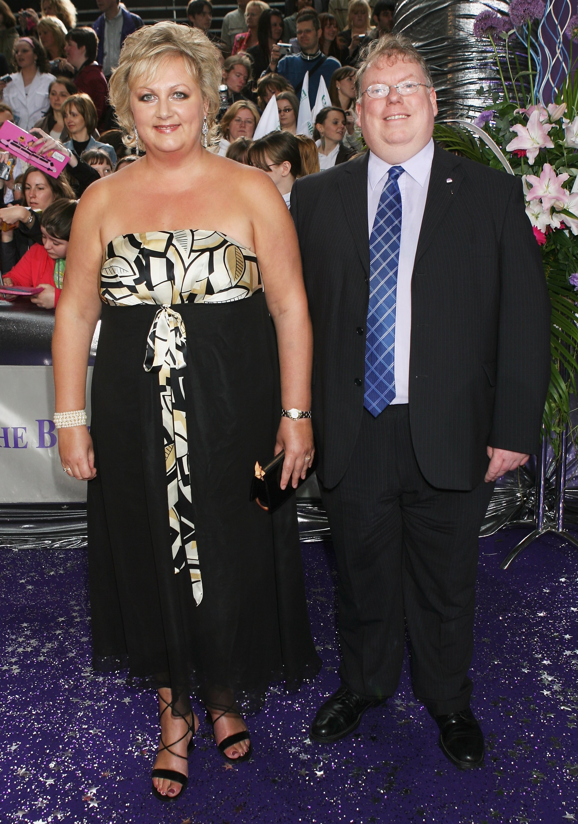 Cleaver arrives at the British Soap Awards in a strapless black-and-gold printed bodice gown tied at the waist, accompanied by a smartly suited guest on the glittering purple carpet.