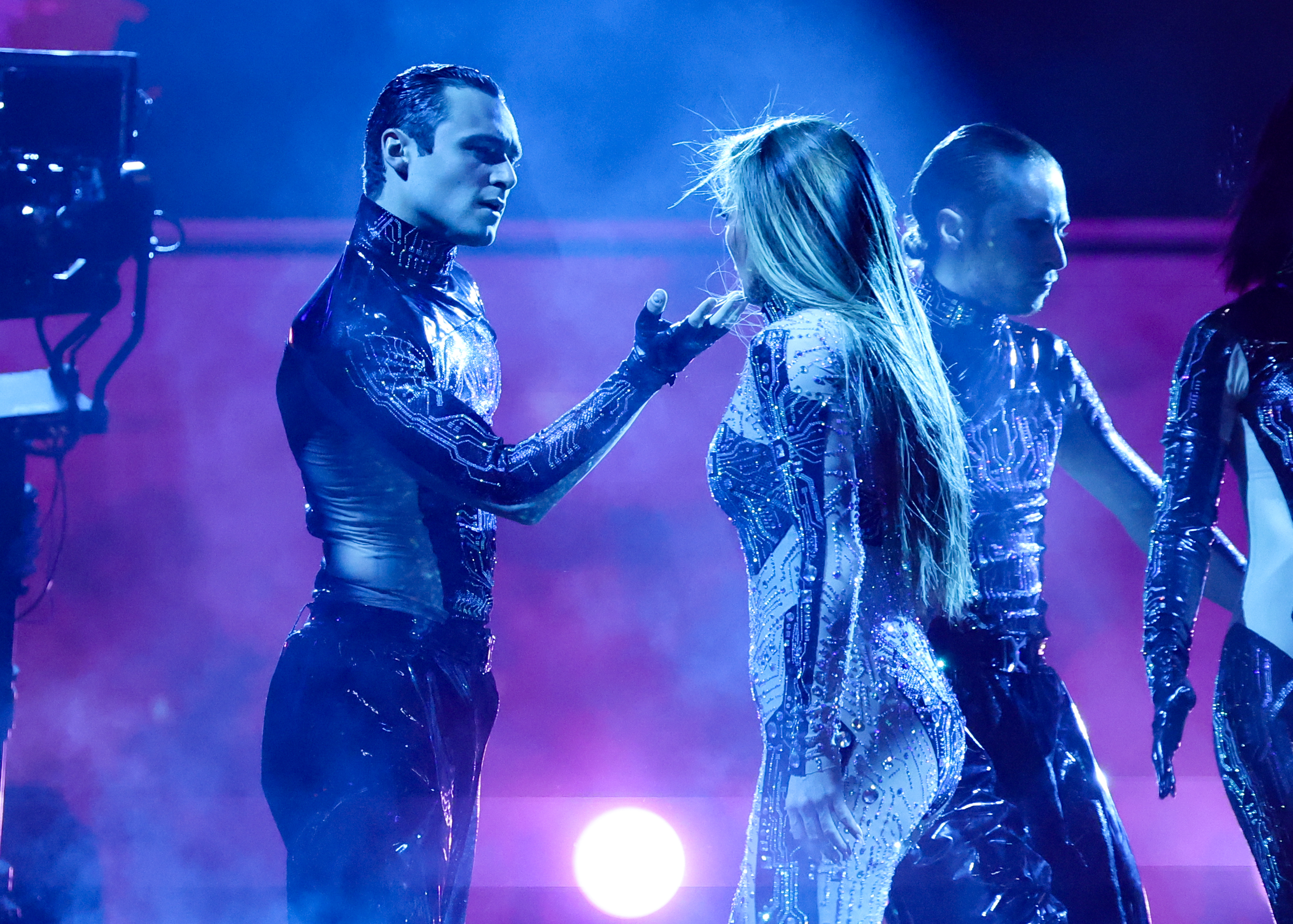 In a fierce and intimate face-off, Jennifer Lopez and a male dancer hold a powerful pose under blue haze, turning her 2025 American Music Awards set at Fontainebleau Las Vegas into a theatrical fusion of dance, tension, and spectacle.