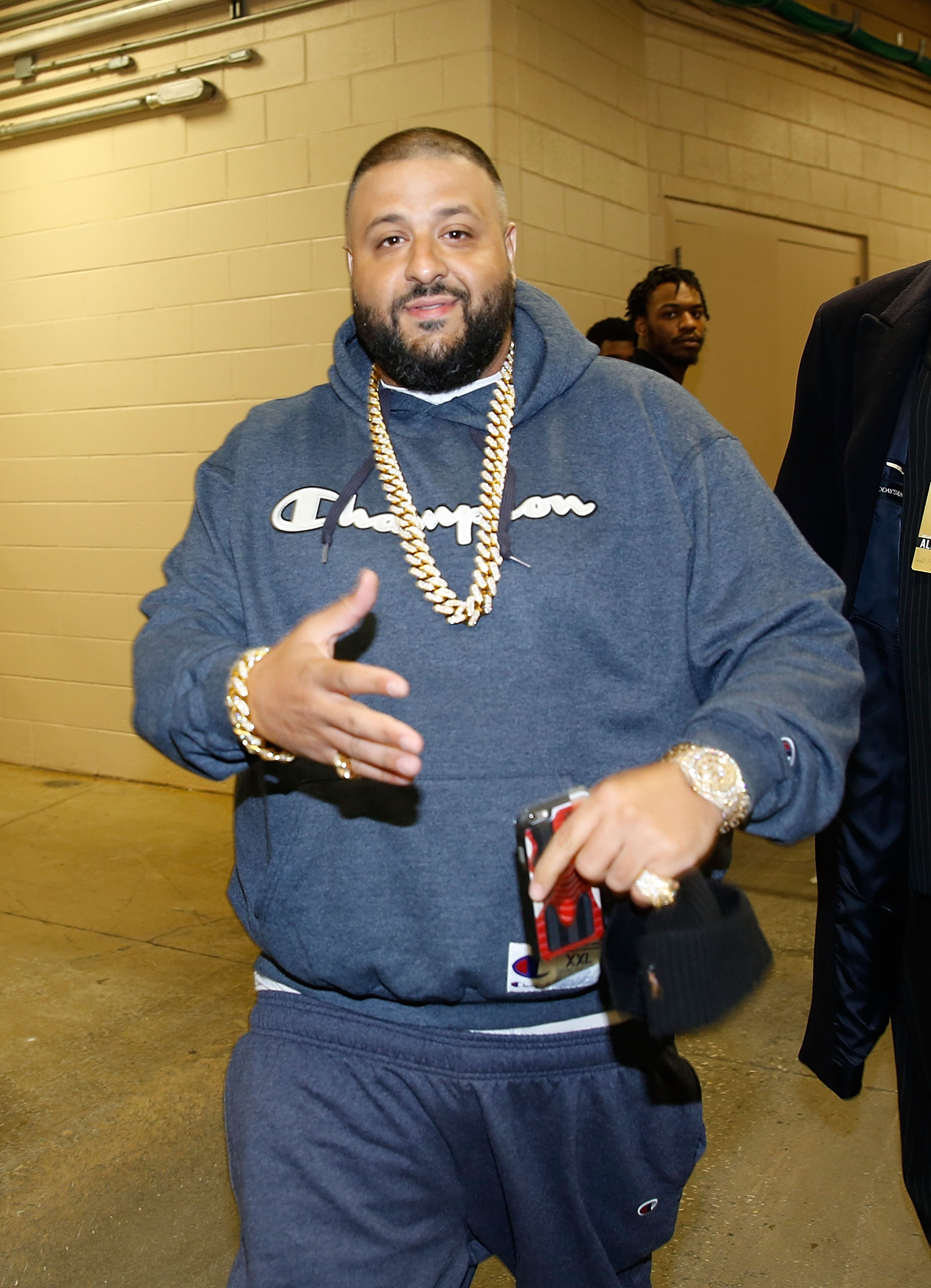 In a grey Champion sweatsuit layered with thick gold chains, DJ Khaled gestures mid-stride while walking through a backstage corridor, accessorized with a statement watch and bracelet that add sparkle to the otherwise relaxed athletic look.