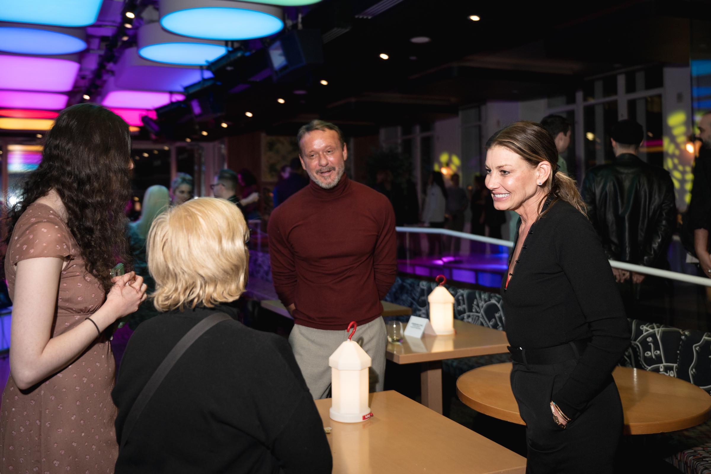 At the opening celebration for Babe at The Green Fig, Faith Hill and Tim McGraw are caught mid-conversation with guests beneath moody lighting. The candid moment feels unguarded, natural, and quietly intimate.