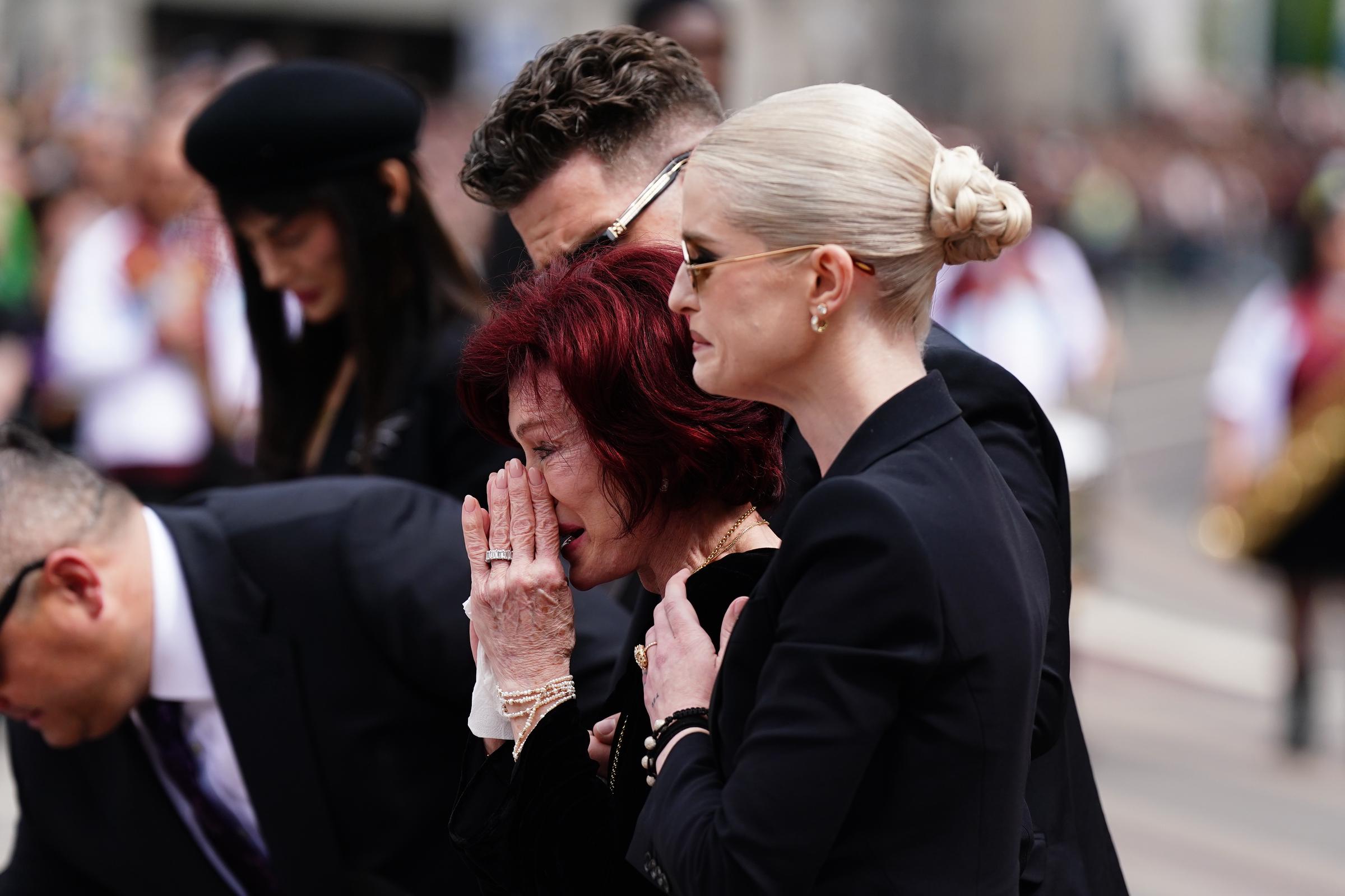 Sharon Osbourne breaks down in tears as her daughter Kelly gently embraces her from behind, offering strength through grief. Surrounded by loved ones, the moment captures a deeply emotional farewell to the rock icon who died on July 22, 2025