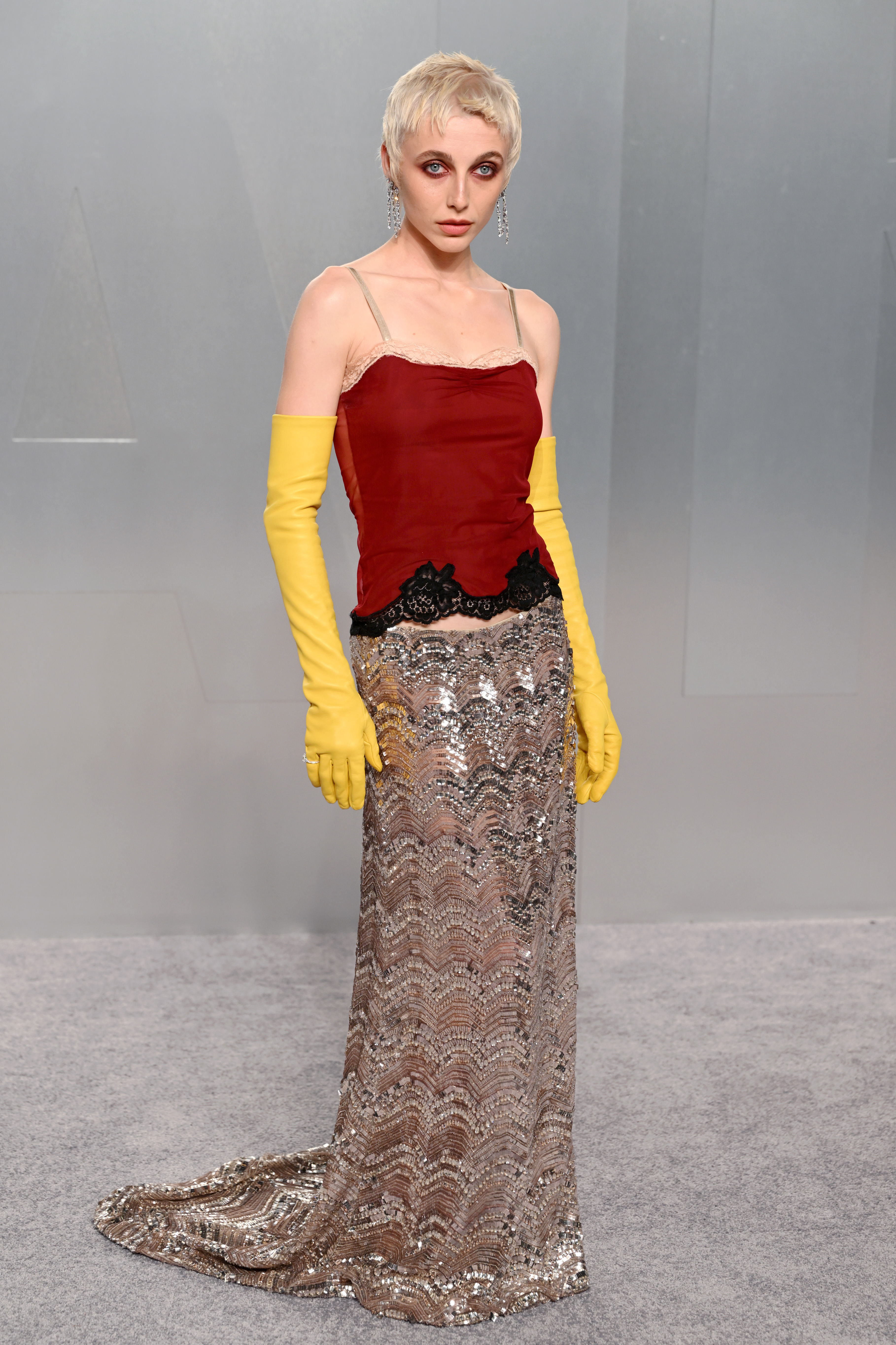 Emma Chamberlain wears a deep red lace-trimmed spaghetti-strap camisole top with black lace detailing at the hem, paired with a floor-length silver sequined chevron-patterned skirt — and bright yellow elbow-length rubber gloves — accessorised with long drop crystal earrings and a cropped platinum pixie cut.