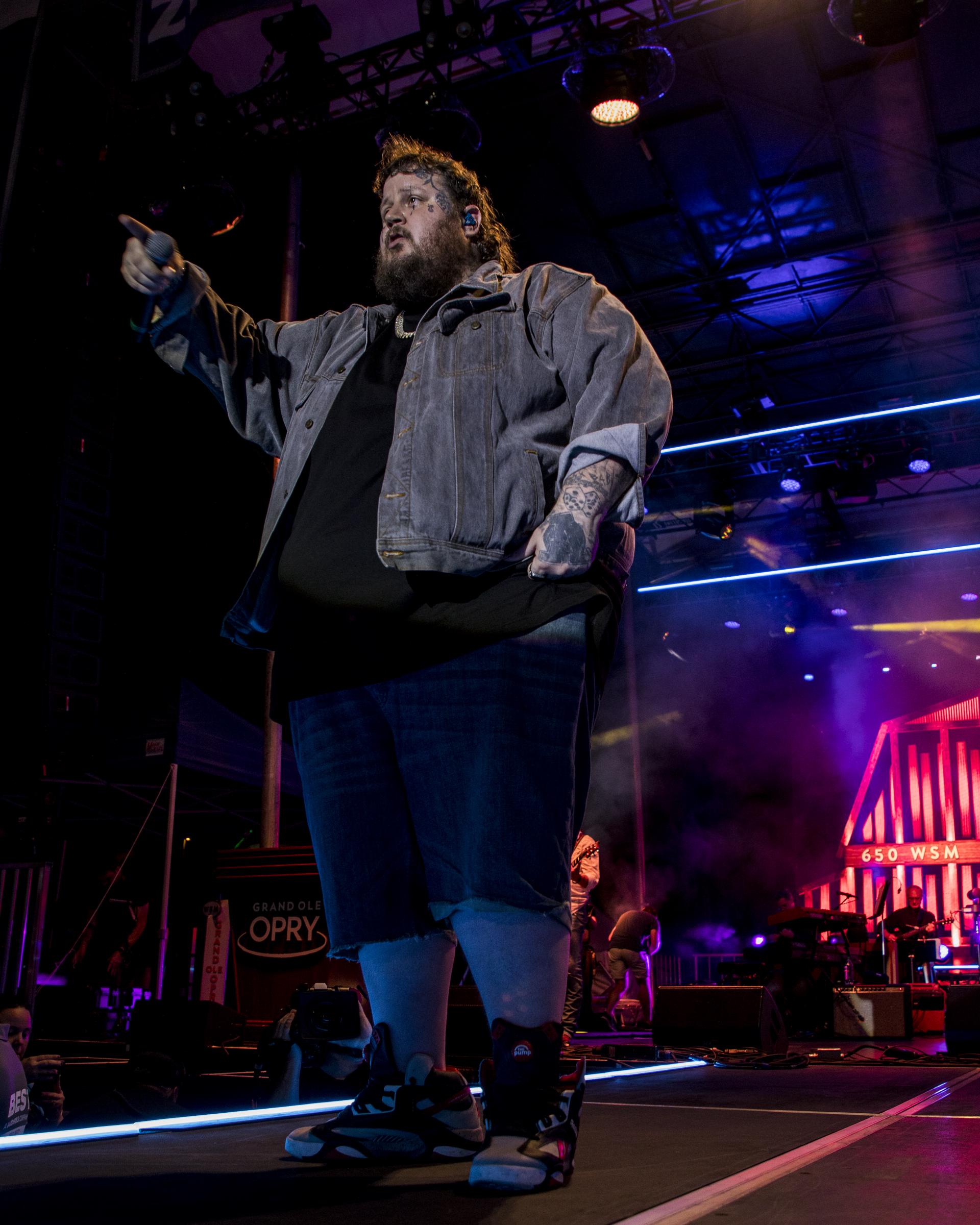 Jason "Jelly Roll" commands the stage. Pointing into the crowd with a powerful and assertive stance, he wears a loose black shirt layered with a faded denim jacket, paired with knee-length denim shorts and black high-top sneakers. The dramatic lighting and colorful stage backdrop amplify his intense presence and connection with the audience.