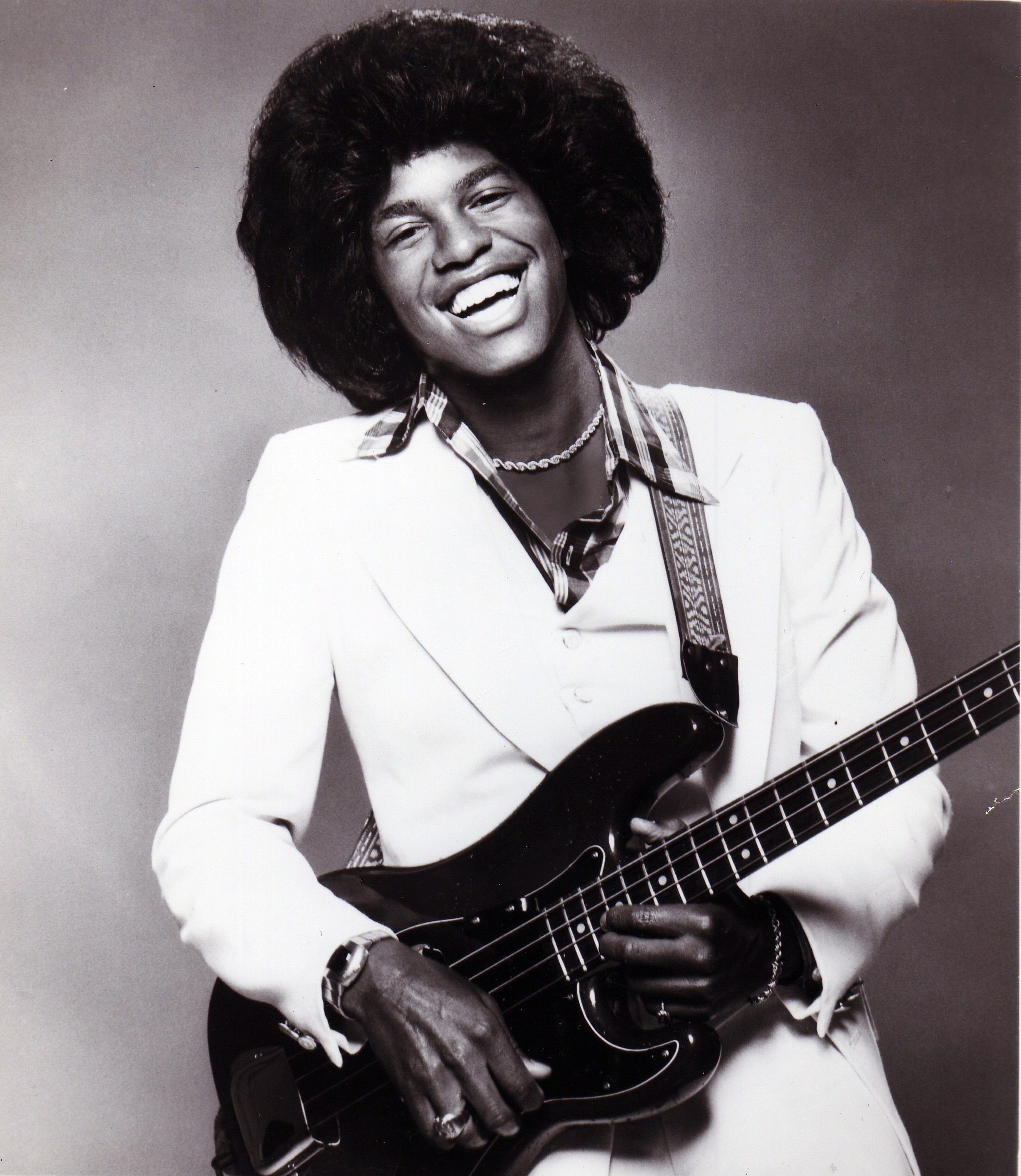Jermaine Jackson poses with a bass guitar, smiling brightly in a crisp white suit layered over a patterned shirt. His voluminous hairstyle and relaxed stance capture the youthful energy and signature style of the era.