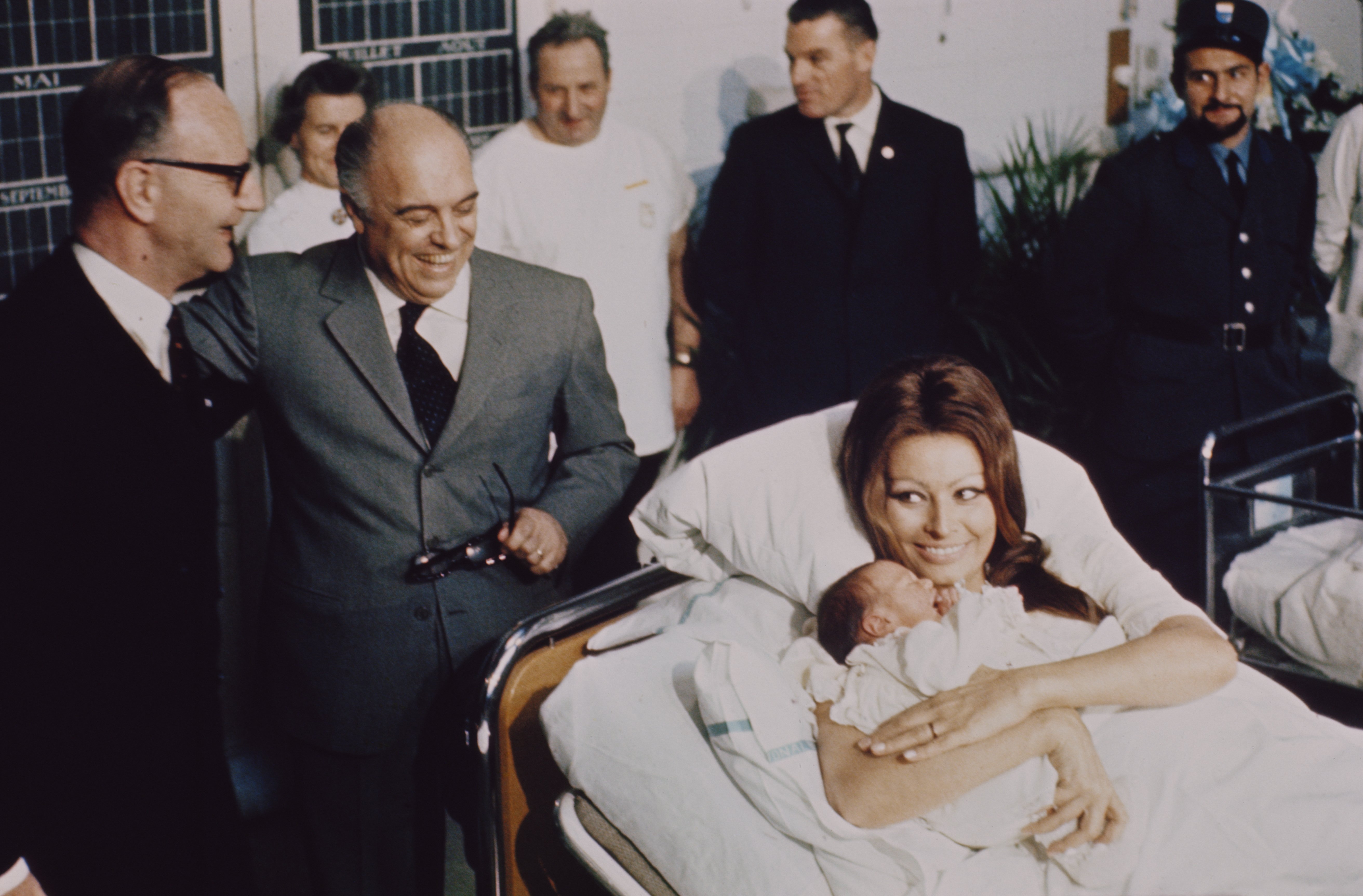 Sophia Loren liegt in einem Krankenhausbett in einer Entbindungsklinik und hält ihren neugeborenen Sohn Carlo junior mit ihrem Ehemann Carlo Ponti, der am 4. Januar 1969 in Genf, Schweiz, neben dem Bett steht. | Quelle: Getty Images