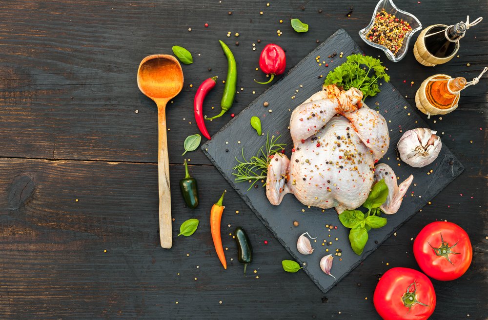 Pollo entero crudo sazonado sobre una tabla, rodeado de ingredientes variados. | Foto: Shutterstock
