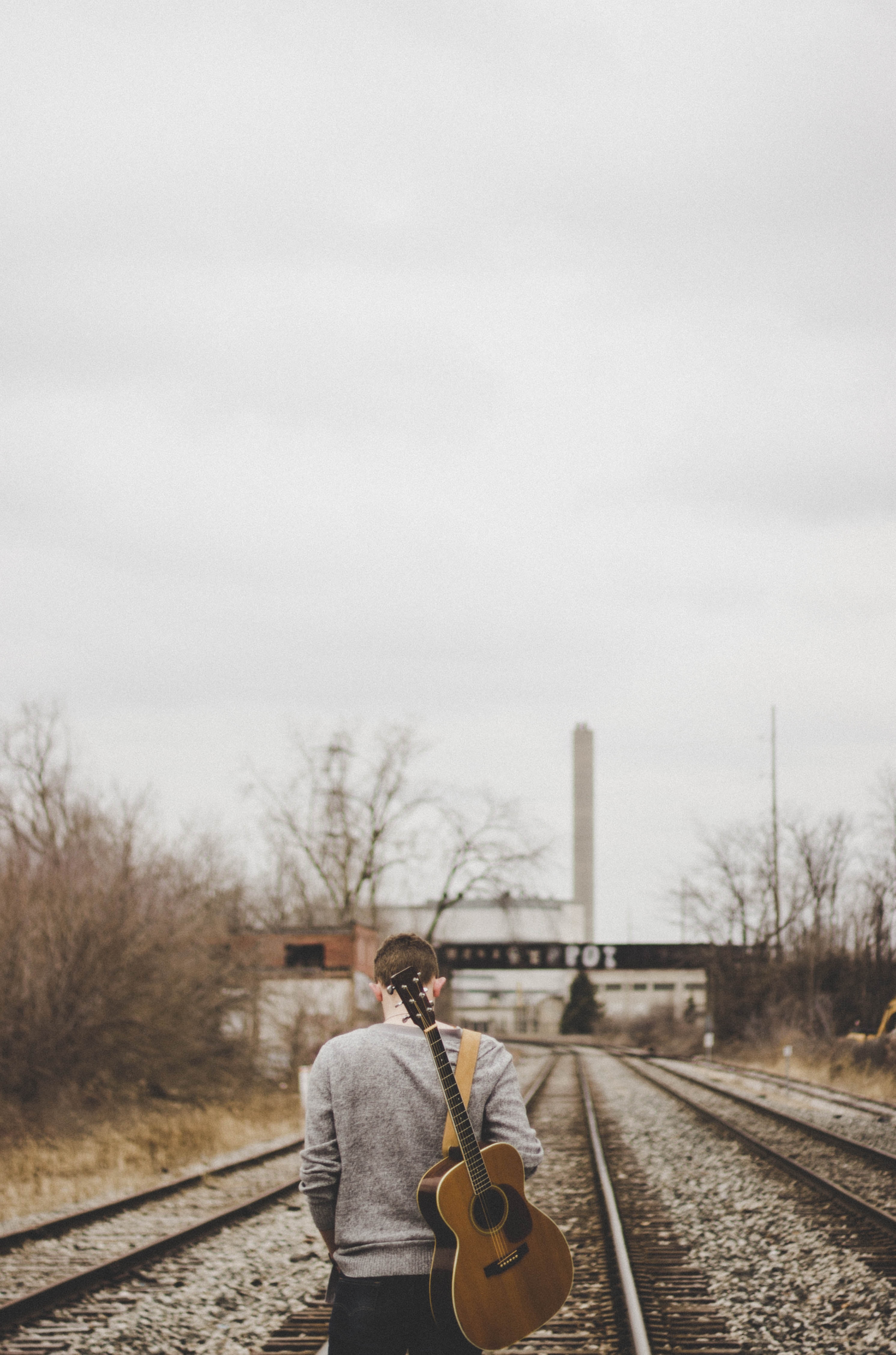 A man walking on a train track while carrying a guitar. │ Source: Pexels