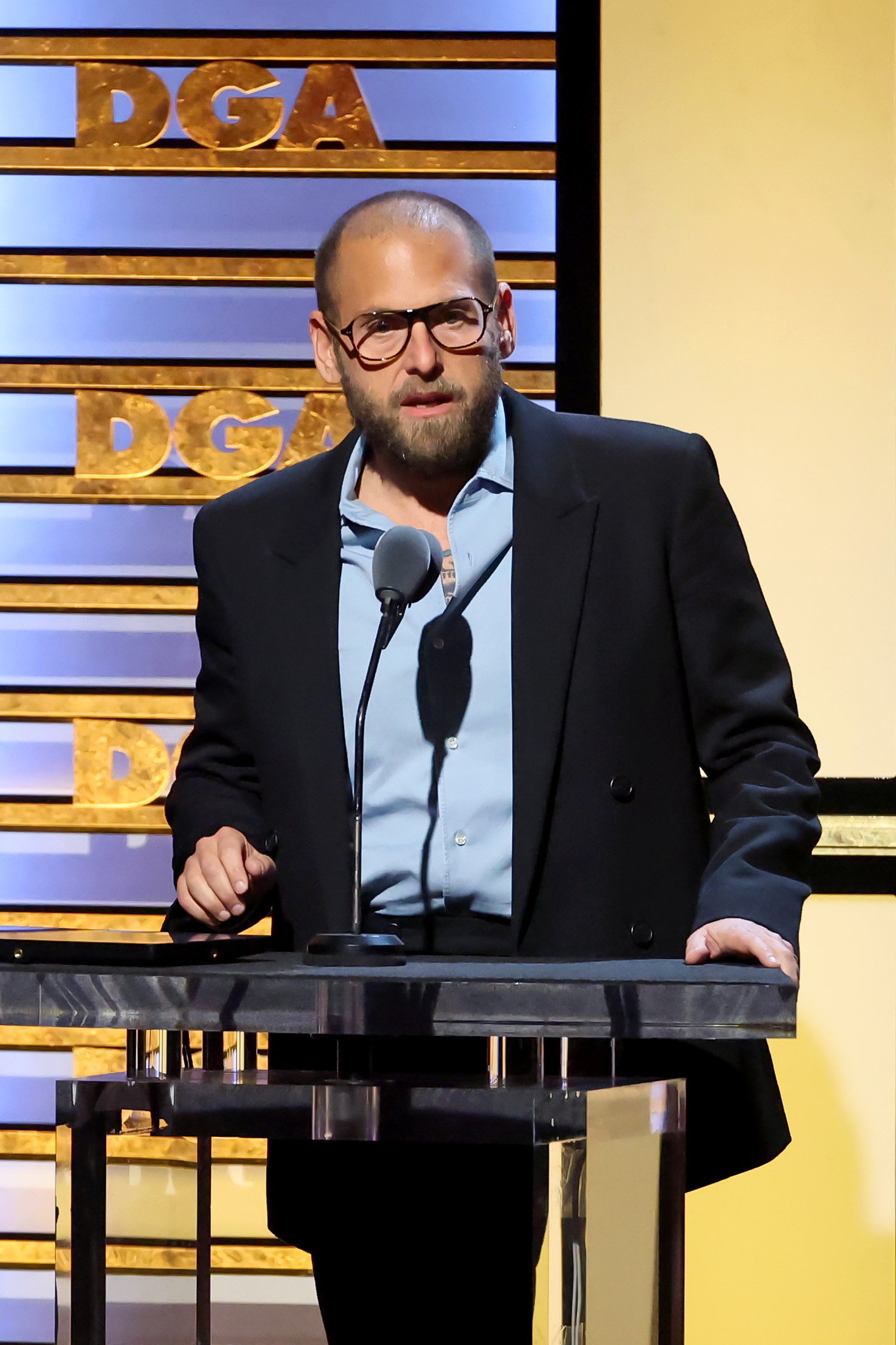 Jonah Hill speaks onstage at the Directors Guild of America Awards, wearing a light blue shirt partially unbuttoned beneath a black double-breasted blazer. Sporting a full beard and oversized glasses, he stands confidently behind the podium with the DGA logo lit up behind him.