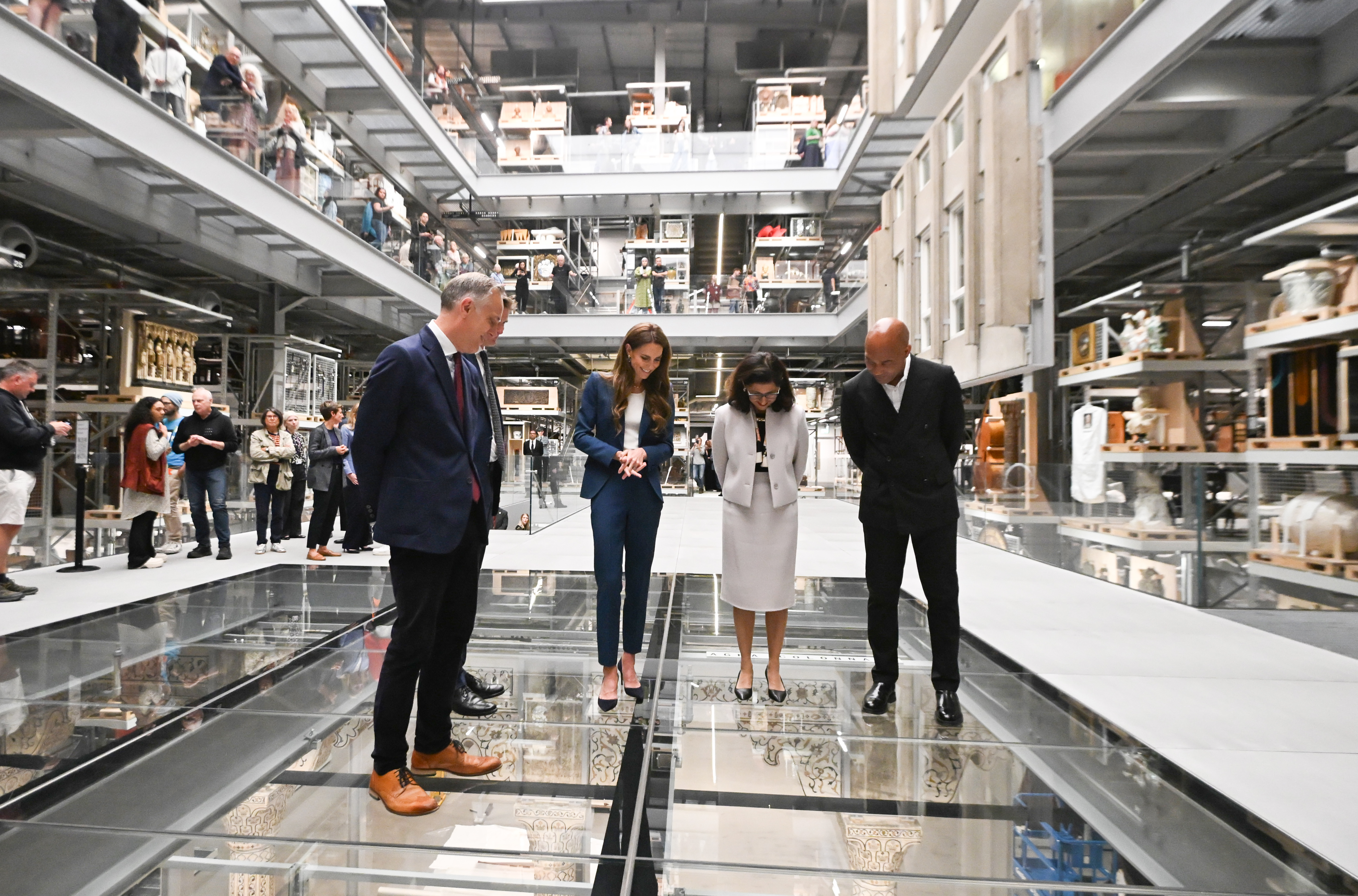 Standing over a glass floor, Princess Catherine observes the display below with fellow museum leaders. Her gaze is downward, posture careful, suggesting both curiosity and caution as they explore the vertical exhibit space.