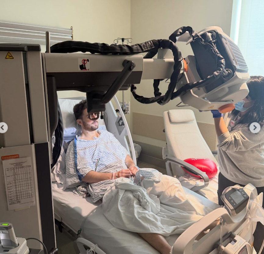 Daniel Neeson lies in a hospital bed wearing a gown and protective glasses as a technician positions a large medical imaging machine above him. | Source: Instagram/bgdans91