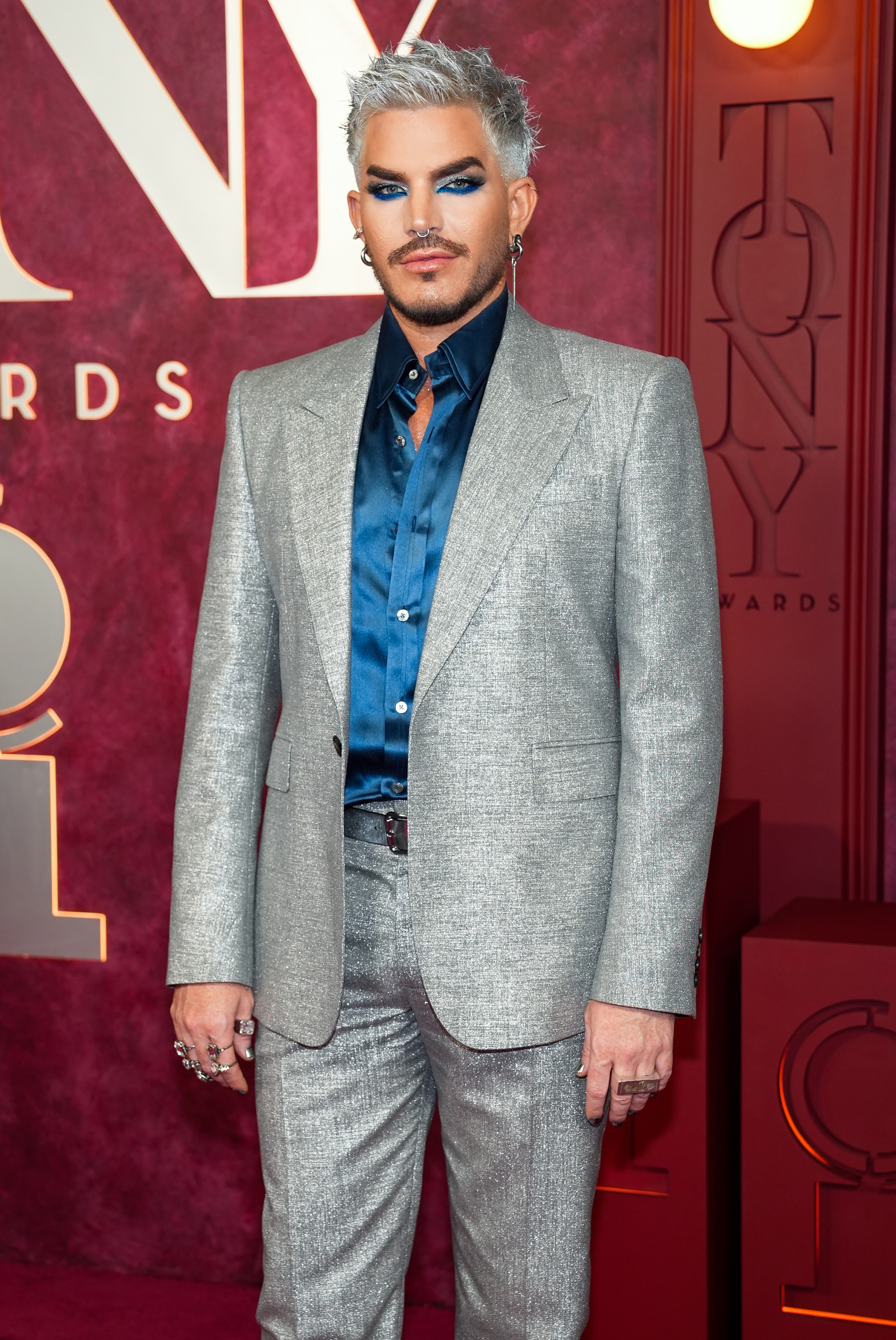 Adam Lambert strikes a composed pose in a textured silver suit layered over a satin blue shirt. His platinum hair and sculpted makeup give the look a polished intensity, while his jewelry adds just the right amount of attitude and individuality.