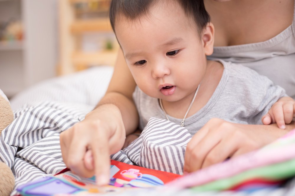 Asian baby looks at book with mom. | Photo: Shutterstock