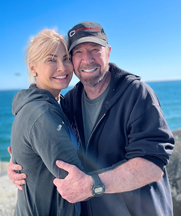Chuck Norris wraps his arm around his wife as they smile brightly by the ocean, the clear blue sky and waves behind them setting a serene scene. | Source: Instagram/chucknorris