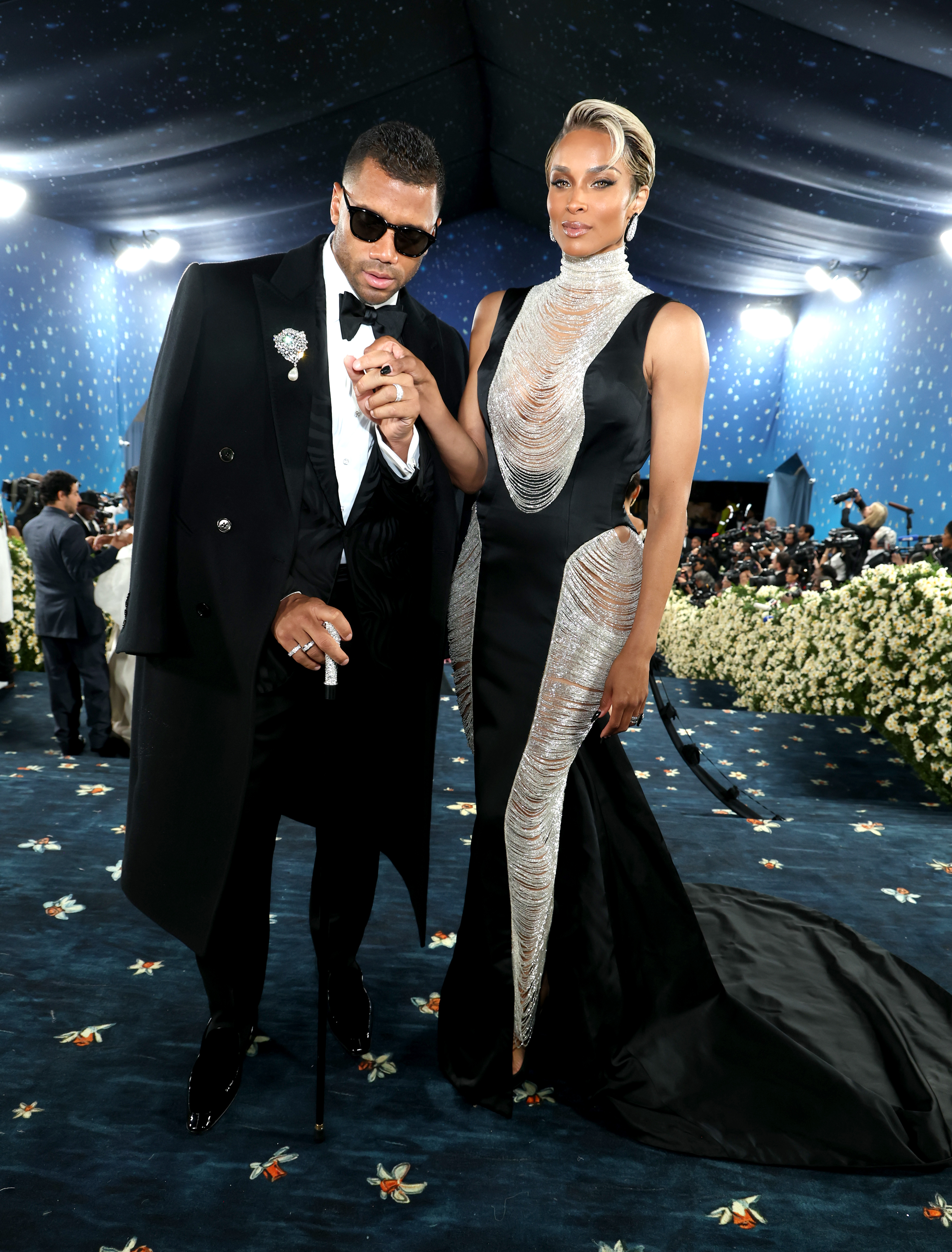 Russell Wilson and Ciara attend the Met Gala at the Metropolitan Museum of Art, embracing the theme with striking, fashion-forward looks. He leans slightly forward with a cane while she stands tall in a detailed, shimmering gown, creating a powerful, editorial-style pose.