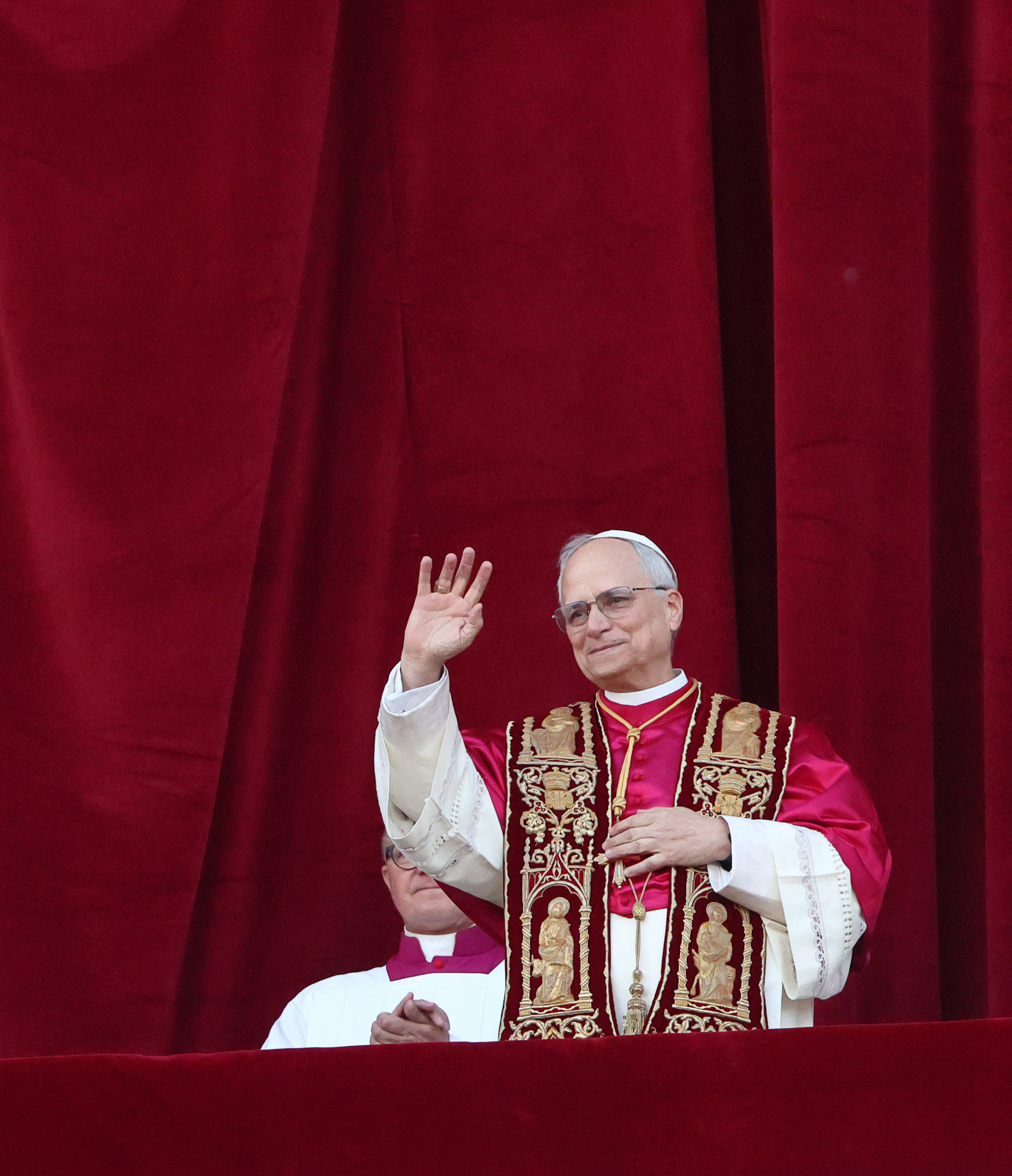 Pope Leo XIV waves softly from the balcony, dressed in deep red vestments adorned with golden iconography, framed by a curtain of rich velvet
