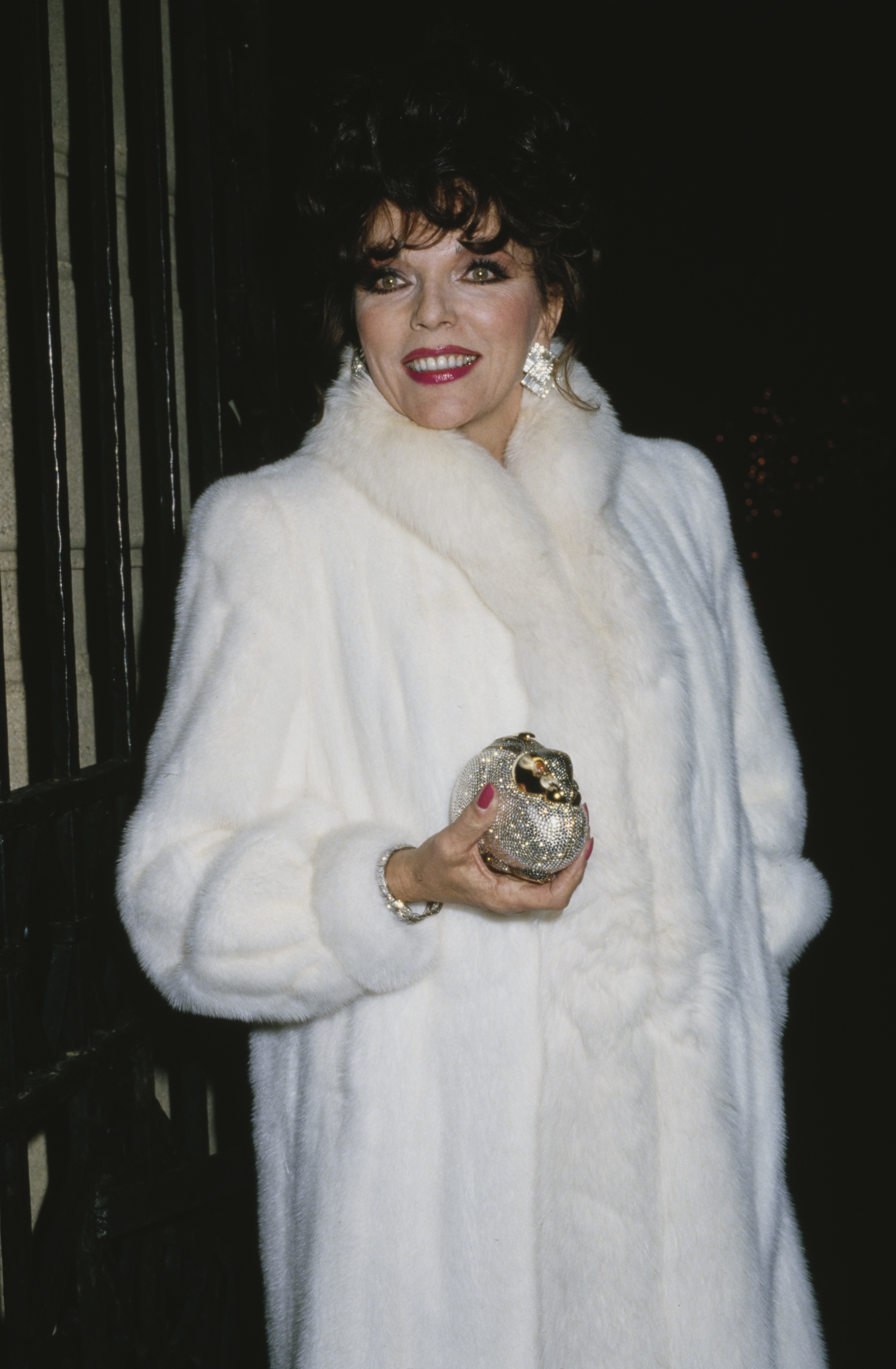 Wrapped in a white fur coat and framed by the glow of flashbulbs, Joan Collins stepped out in Los Angeles, California, circa 1980s, embodying the unapologetic opulence of the decade. With her crystal-studded clutch, statement earrings, and trademark glamour, she exuded the polished confidence that mirrored her defining role on primetime television during the era.