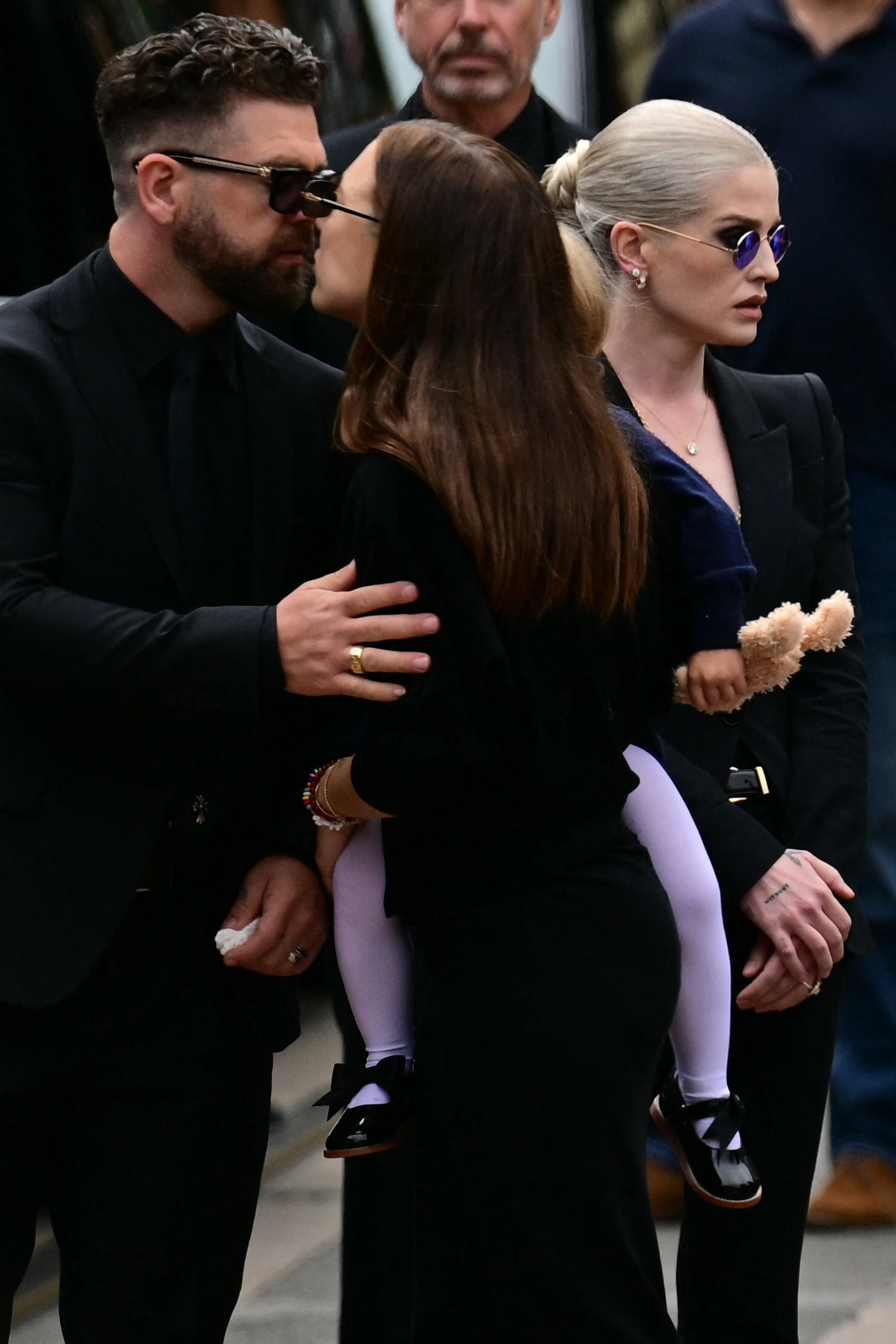 Jack Osbourne kisses his wife Aree as she holds their child while sister Kelly stands by them at the Black Sabbath Bridge in Birmingham, England on July 30, 2025 during their late father's funeral procession. | Source: Getty Images