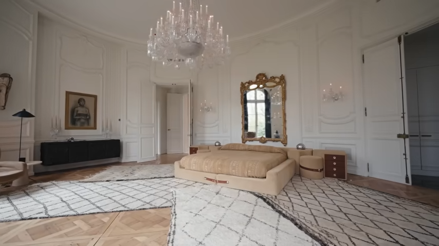 The majestic, oval-shaped master bedroom features pristine white wood moldings, a grand crystal chandelier, and a custom tan suede platform bed. | Source: YouTube/@Archdigest