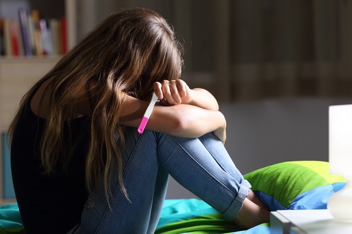 Vista lateral de una adolescente embarazada triste sentada en su cama después de revisar una prueba de embarazo | Foto: Shutterstock.com