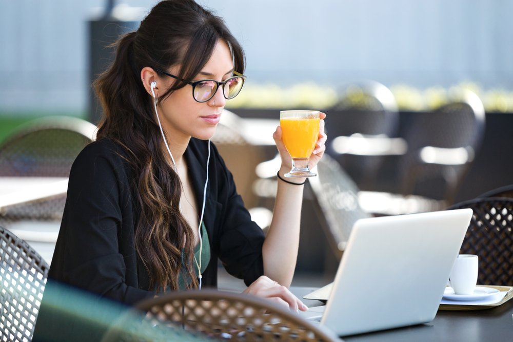 Aufnahme einer jungen Frau, die Orangensaft trinkt, während sie mit ihrem Laptop in einem Café arbeitet. I Quelle: Shutterstock