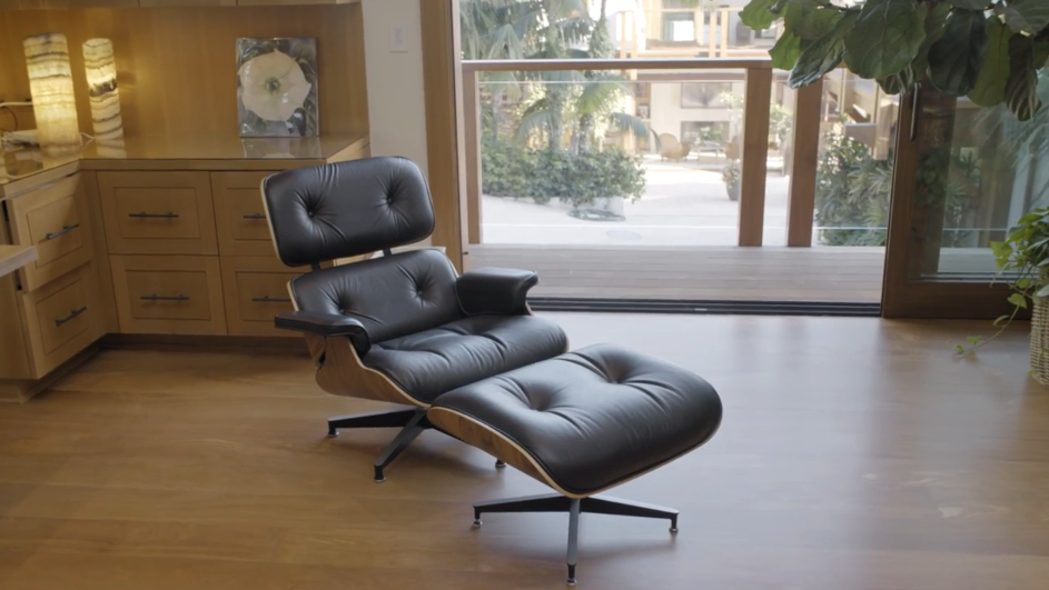 A black leather Eames lounge chair and matching ottoman sit on polished hardwood floors in Pierce Brosnan's office space. In the background, built-in light wood desk cabinetry and soft glowing lamps complement large glass sliding doors that open out onto a sunlit balcony overlooking the courtyard. | Source: YouTube/Architectural Digest