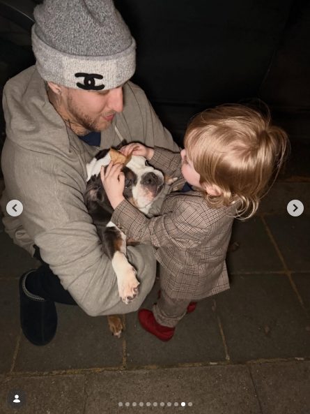 A second shot from the same pavement moment, taken from a slightly different angle, with Langford cradling the bulldog on his knee while the toddler stretched both arms out to hold the animal. Shot from above, the three-year-old's red shoes are the brightest detail in an otherwise dark frame. | Source: Instagram/kellyosbourne