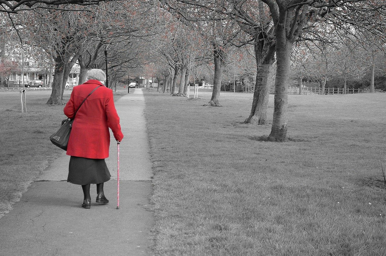 An elderly woman wearing a red jacket and walking with a cane at a park  | Source: Pixabay