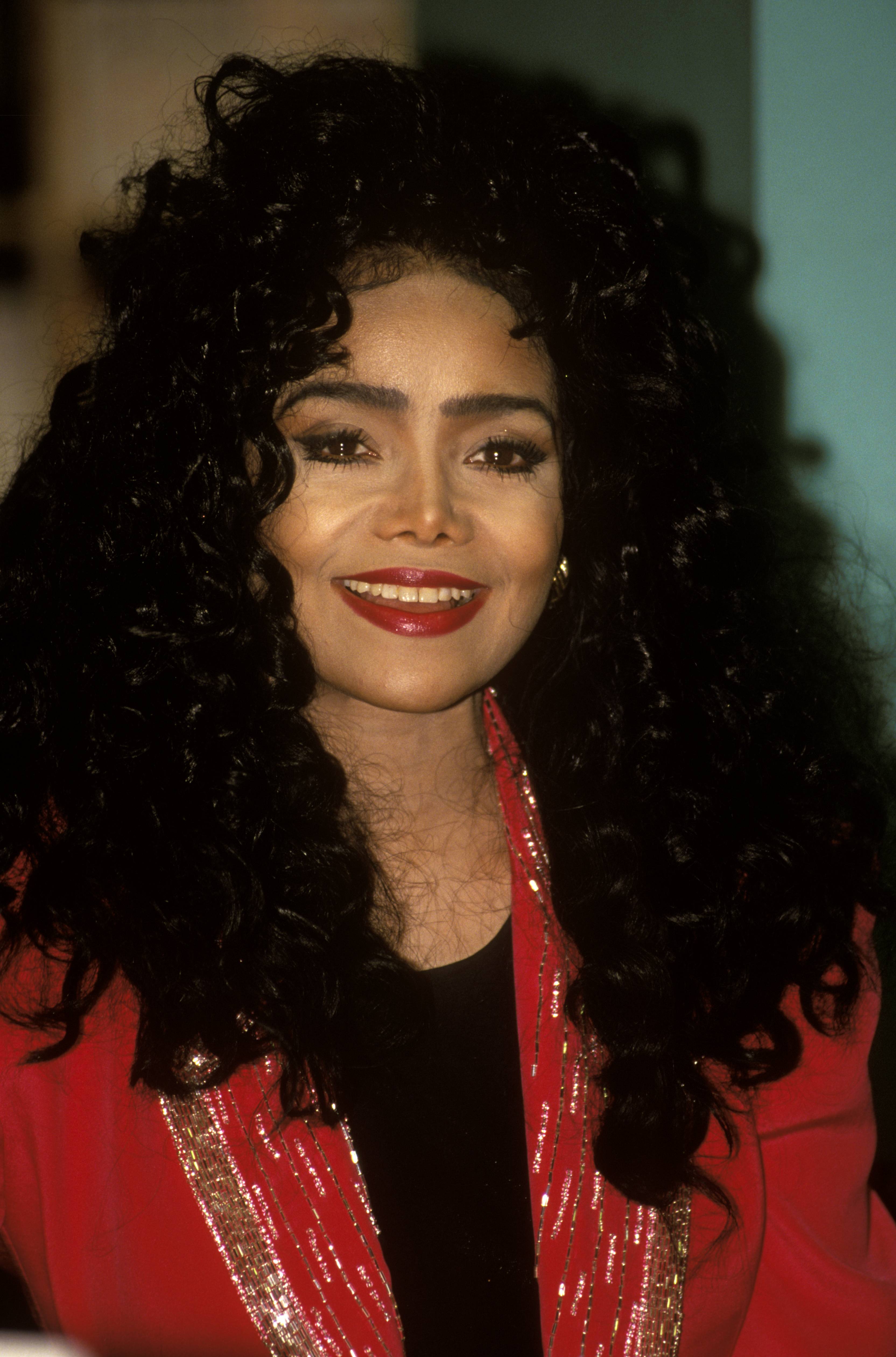 Wearing a red sequined-trim jacket and signature voluminous curls, La Toya Jackson flashes a confident smile. Her bold makeup and radiant style are unmistakably her trademark during her '90s public appearances.