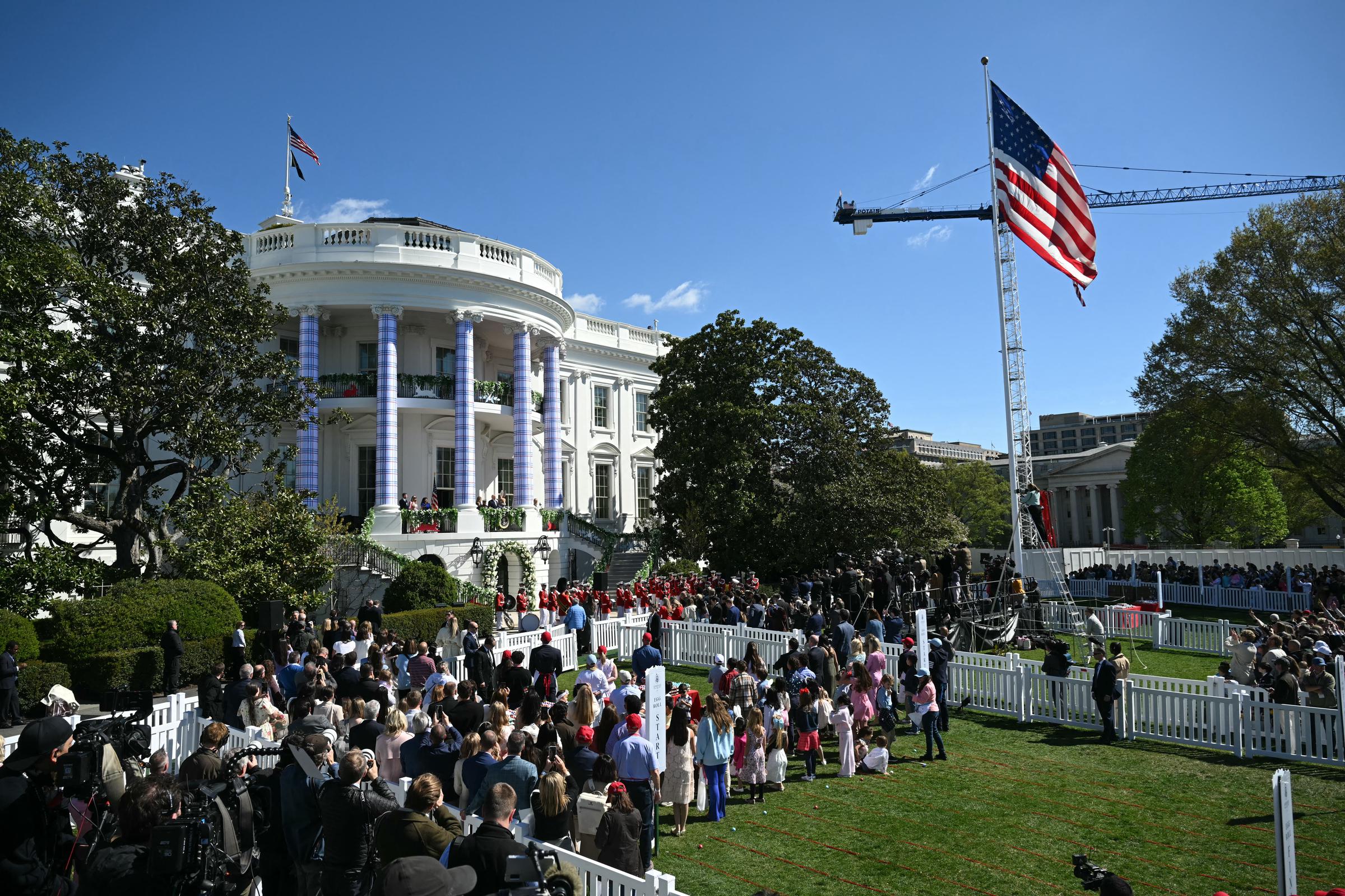 Donald and Melania Trump appear before a crowd on the South Lawn as guests gather for the 2026 White House Easter Egg Roll, with the decorated balcony and a large American flag underscoring the festive setting.