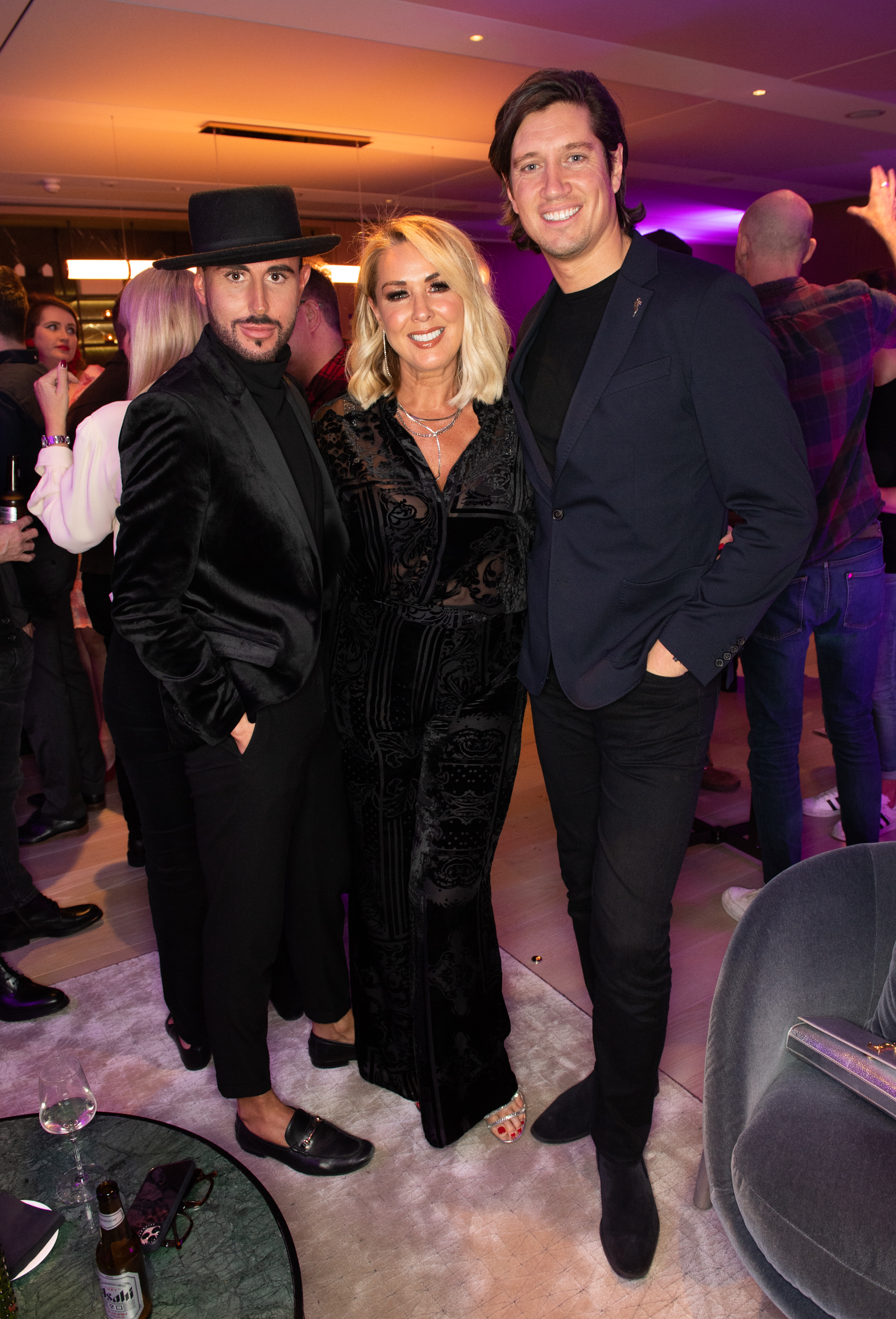 Vernon Kay, in a dark blazer and black trousers, smiles beside Claire Sweeney, who stuns in a sheer black outfit, as they pose together with company at a lively evening event.