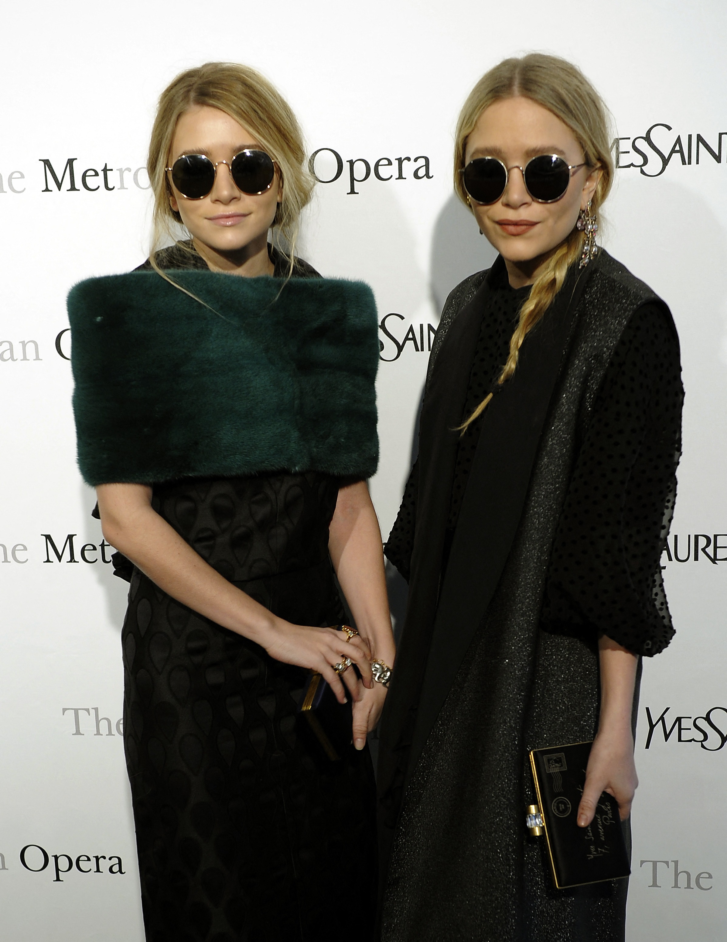 Mary-Kate and Ashley Olsen stand side by side at a formal event, both in dark outfits and round sunglasses. Their composed expressions and sleek styling create a bold, modern look.
